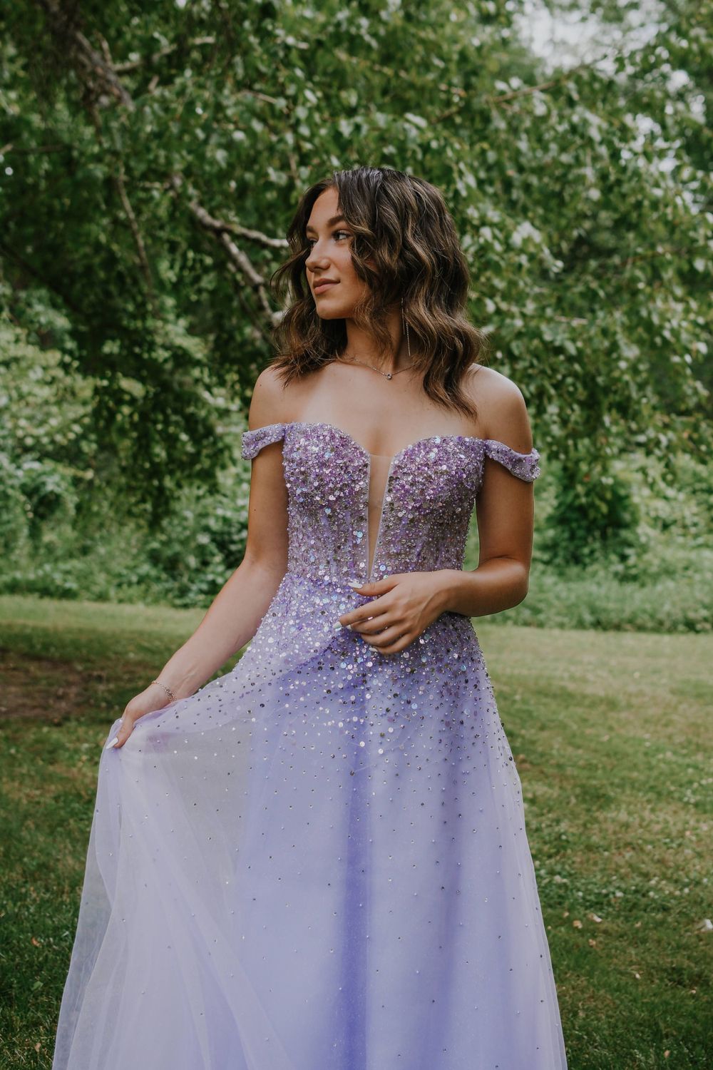 A lavender off-shoulder prom gown with beaded bodice and flowing tulle skirt sparkles in an outdoor garden setting.