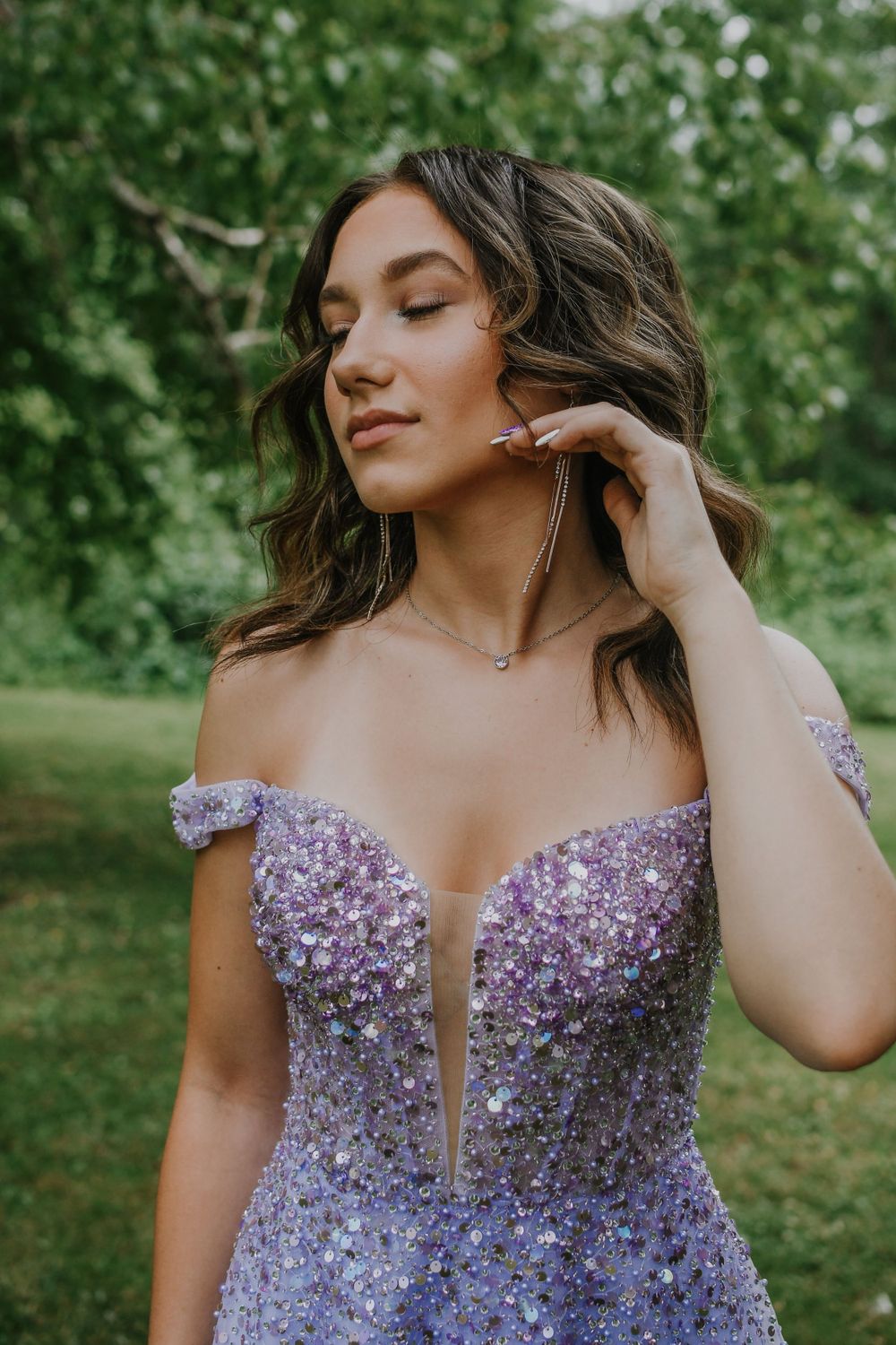 Elegant purple sequined prom dress with off-shoulder design shown in outdoor garden setting with green foliage background.