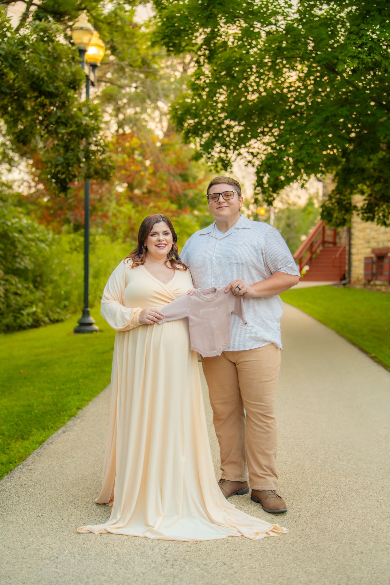 A romantic outdoor couple portrait on a path with green trees and lampposts in warm evening light.