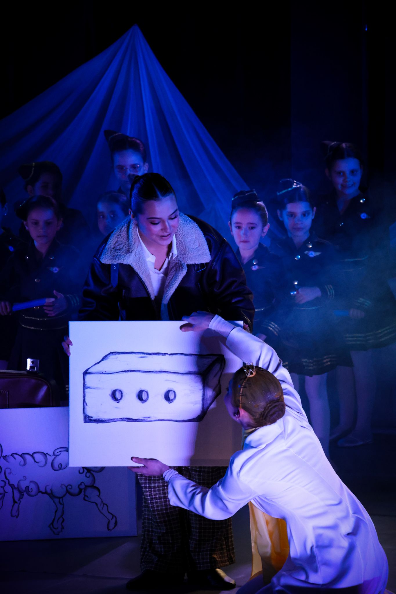 Theater scene with performers interacting with illuminated prop boxes in blue lighting.