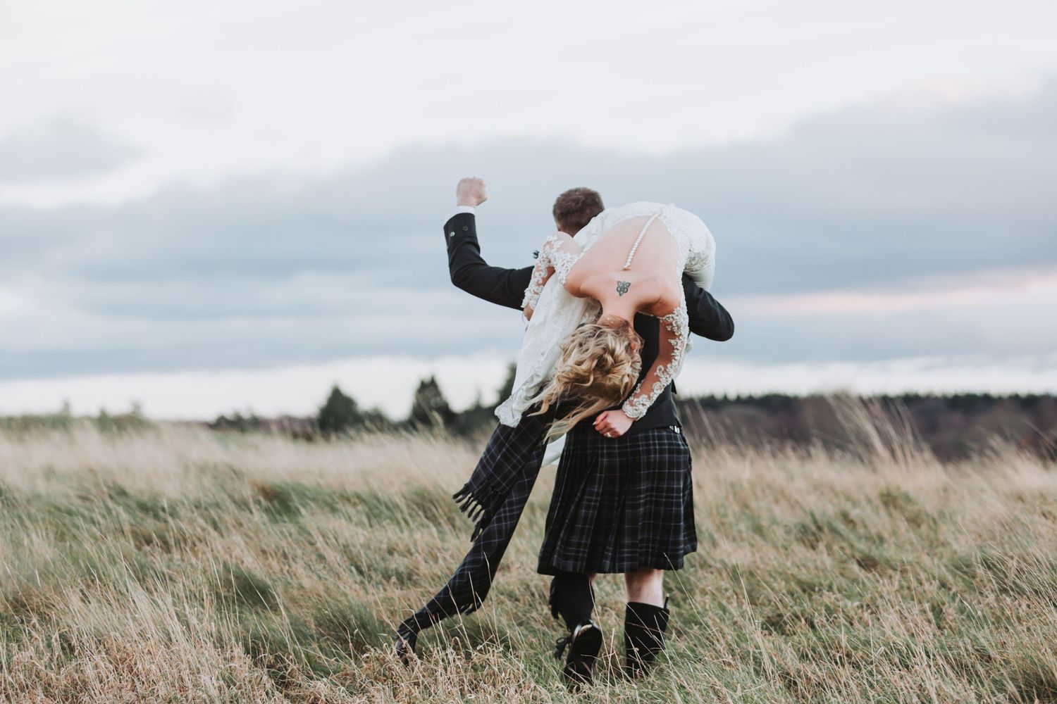 A groom in a kilt joyfully lifting his bride in a sweeping field under a dramatic cloudy sky.