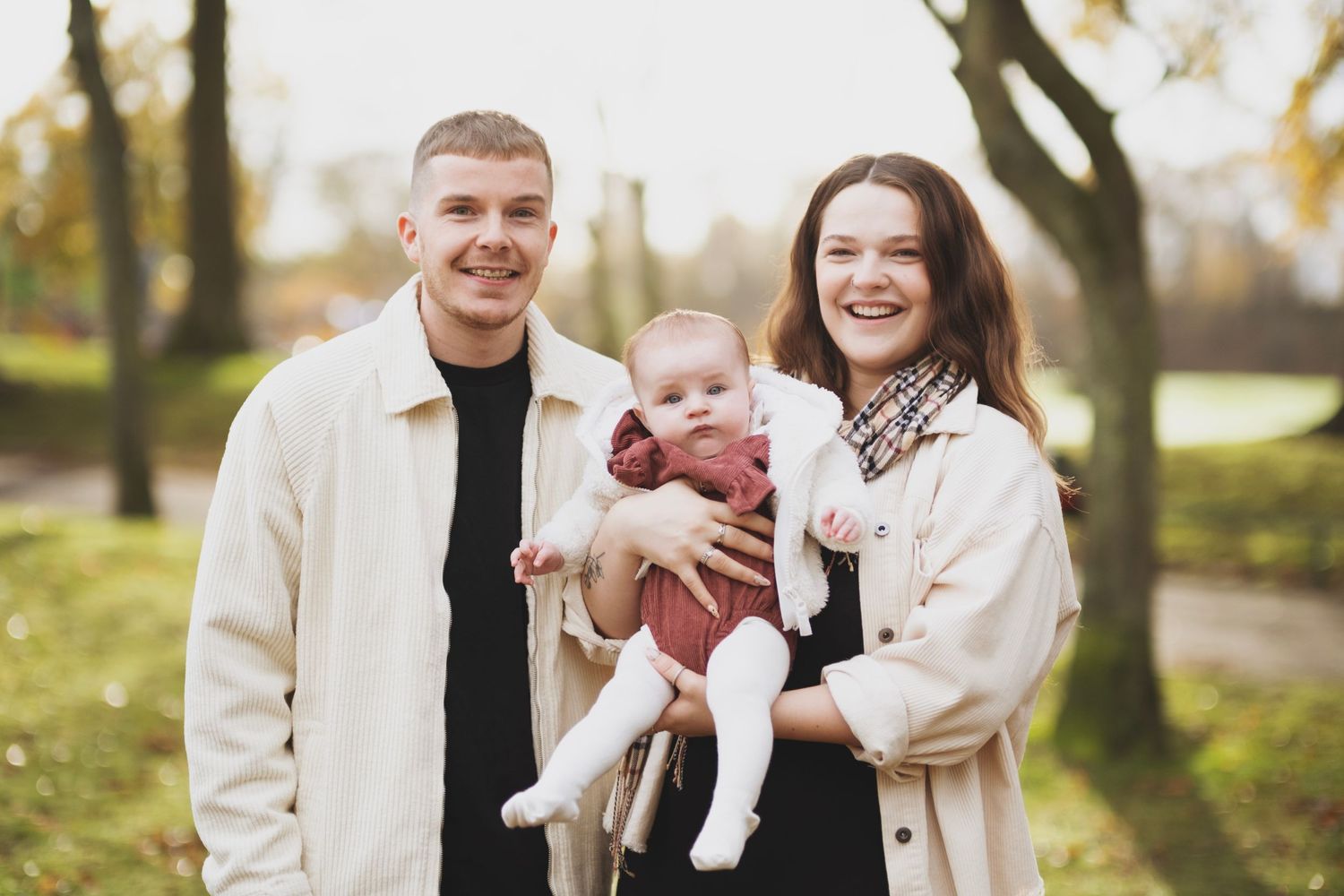Young couple smiling in autumn park holding their baby dressed in red.