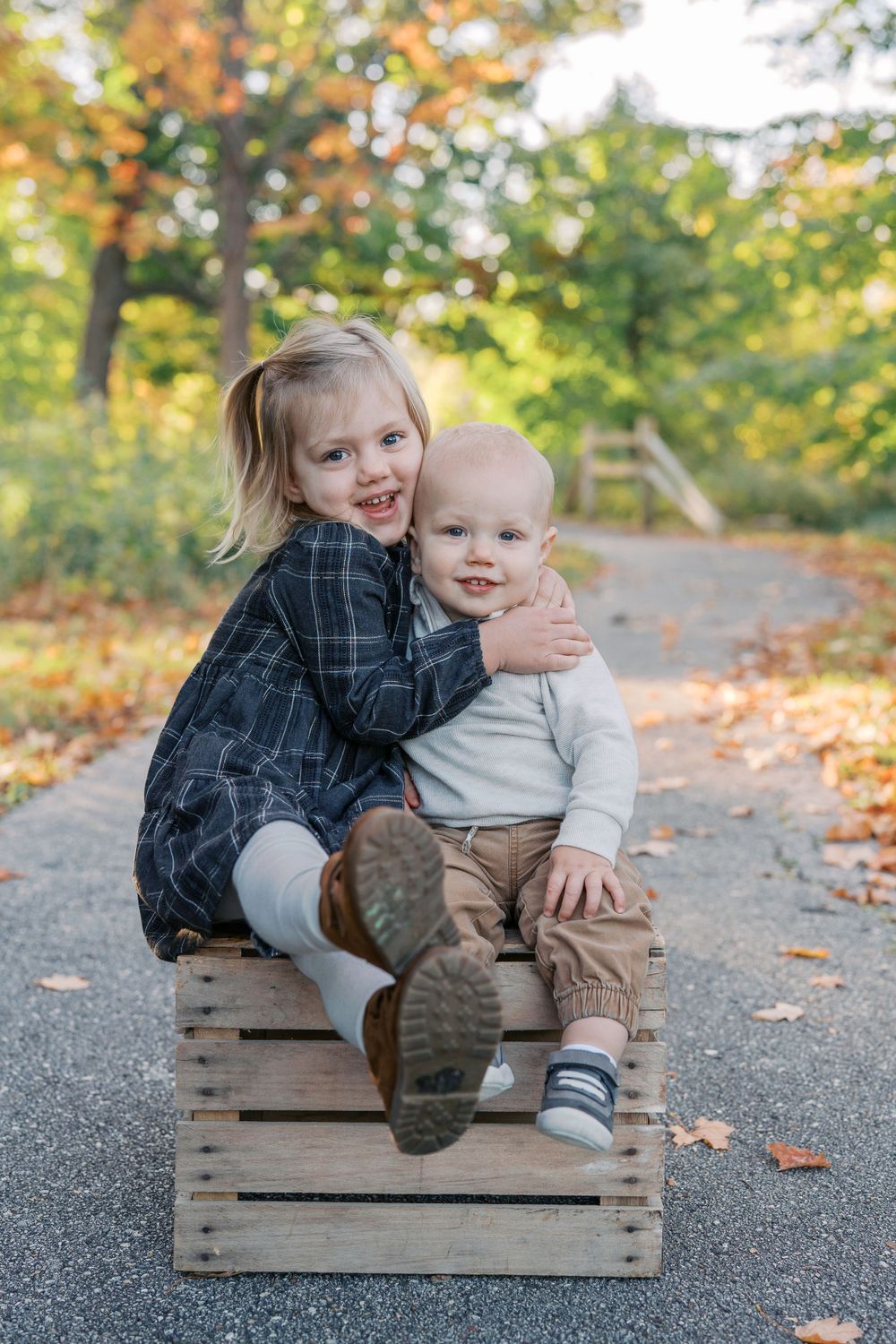 Siblings sitting on a wooden crate during an autumn outdoor photoshoot.
