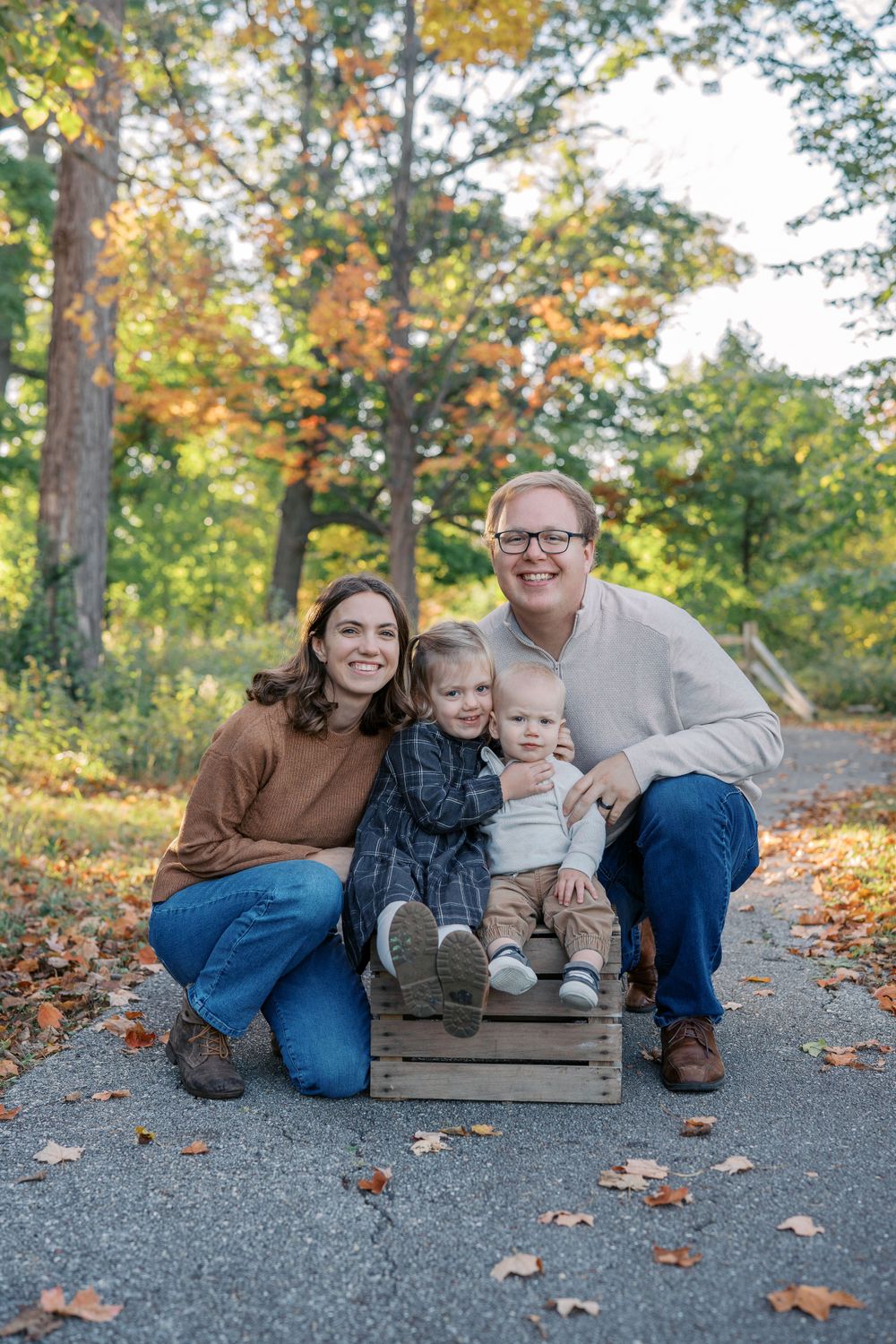 Family relaxing on wooden crates during a fall photo session on a gravel path.