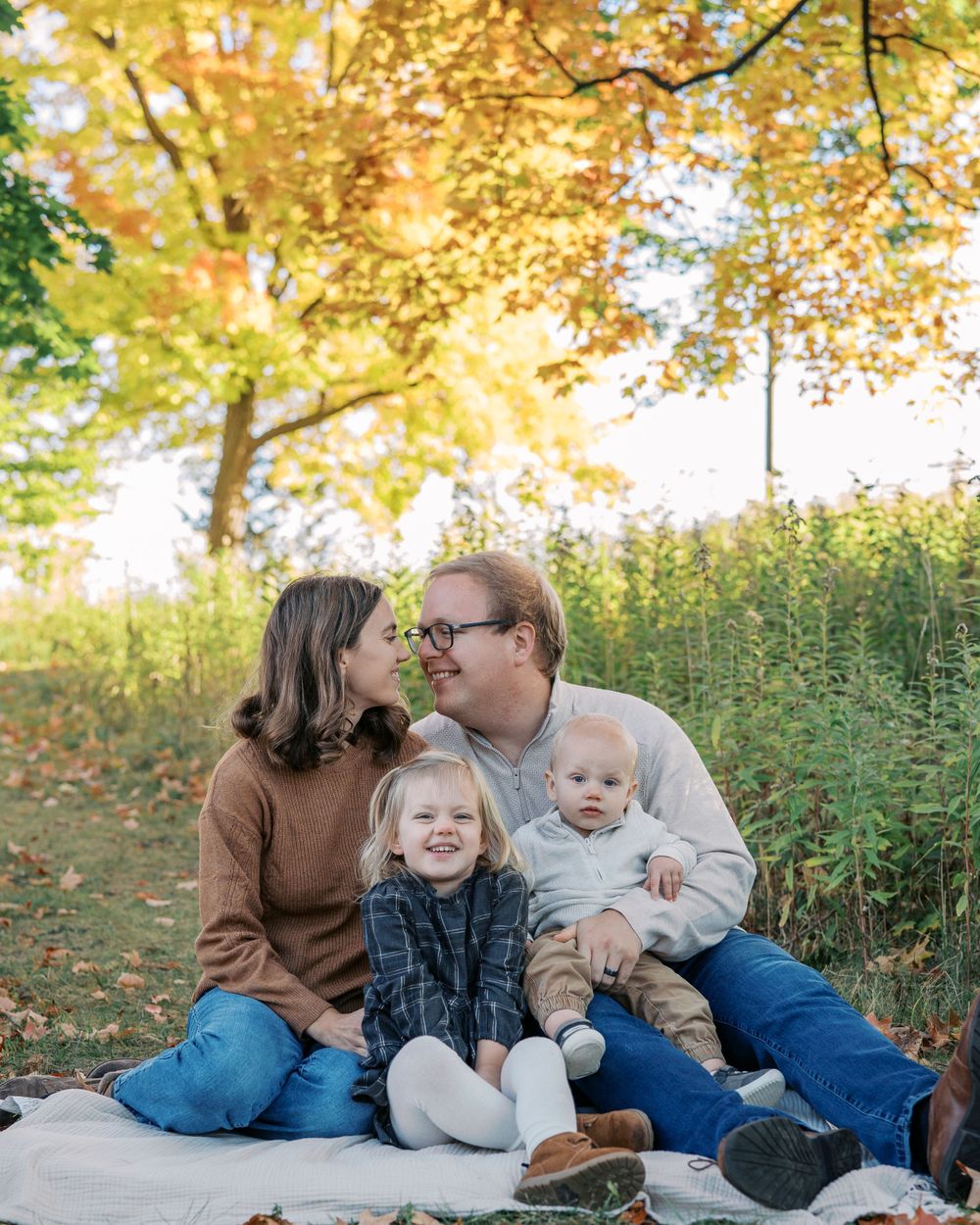 Family enjoying autumn picnic on blanket under golden fall foliage in park setting.