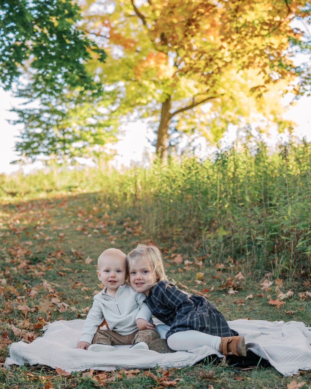 Two small children sitting together on white blanket during fall photo session.