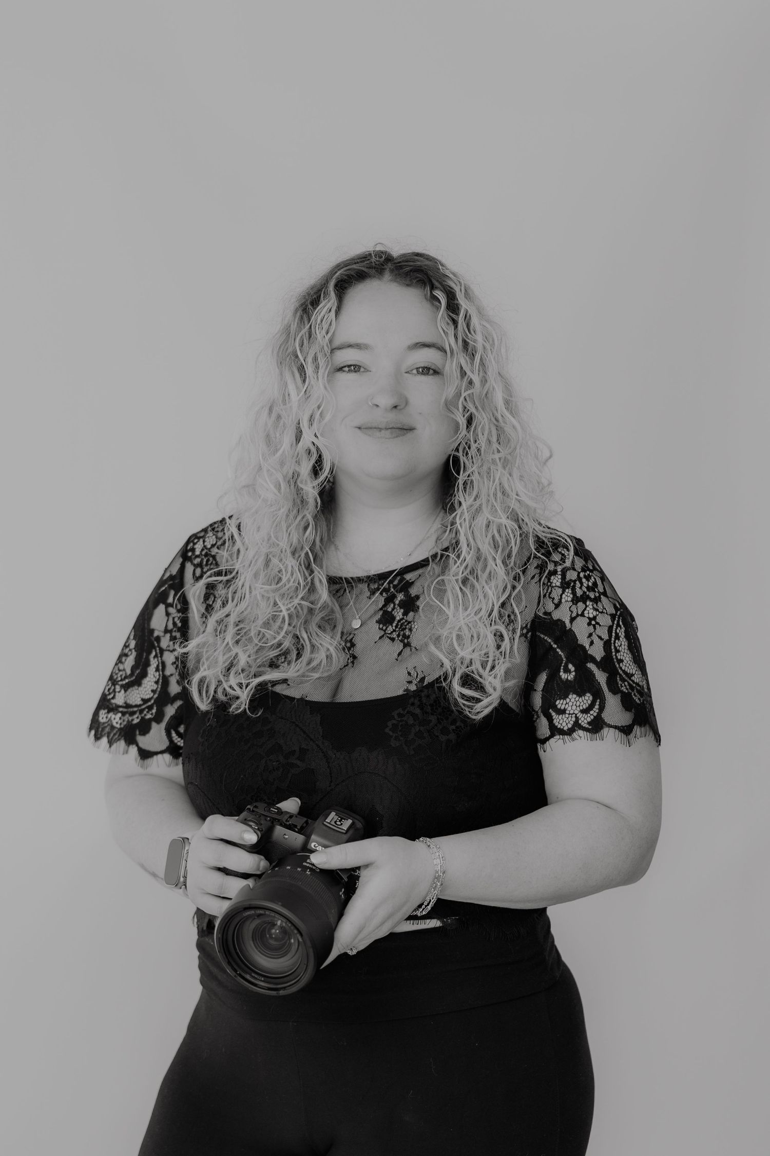 Photographer in black lace dress holding camera against grey background.