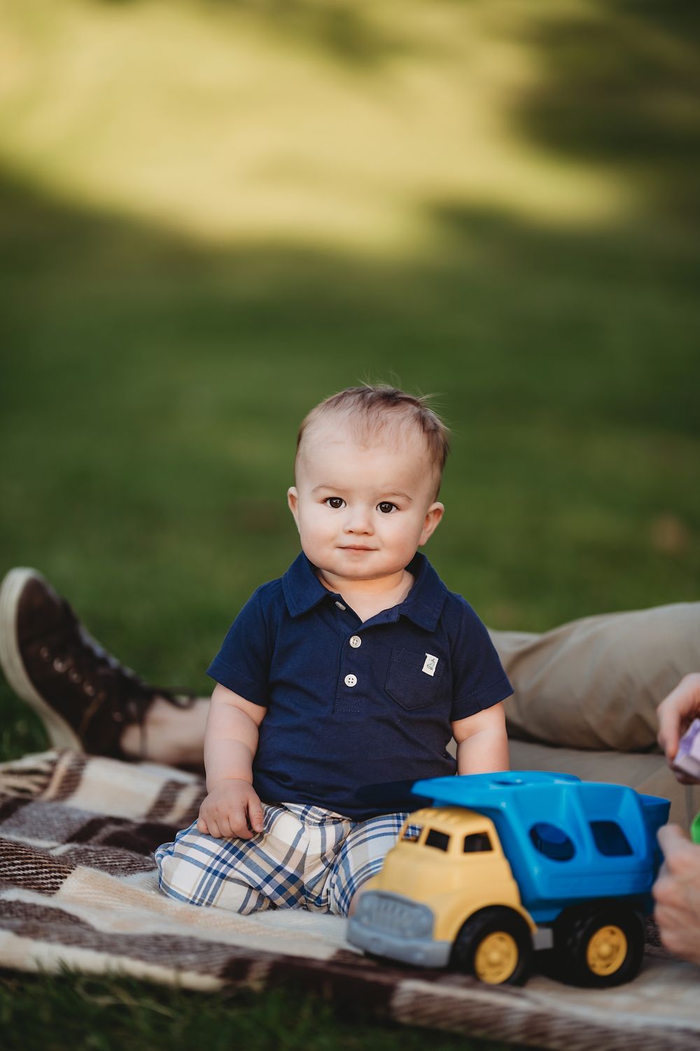 A small child sits on a blanket outdoors wearing a navy polo shirt with a yellow toy dump truck nearby.