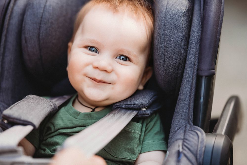 A smiling infant wearing a green shirt sits securely strapped in a gray car seat or stroller.