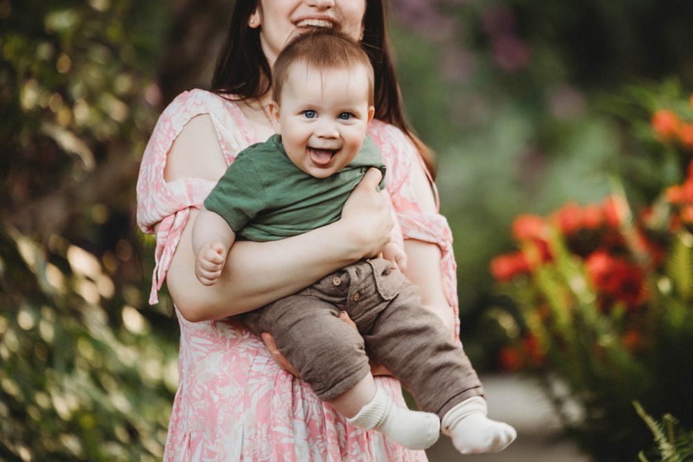 Smiling baby in green shirt and tan pants is held up outdoors against blurred floral garden background.