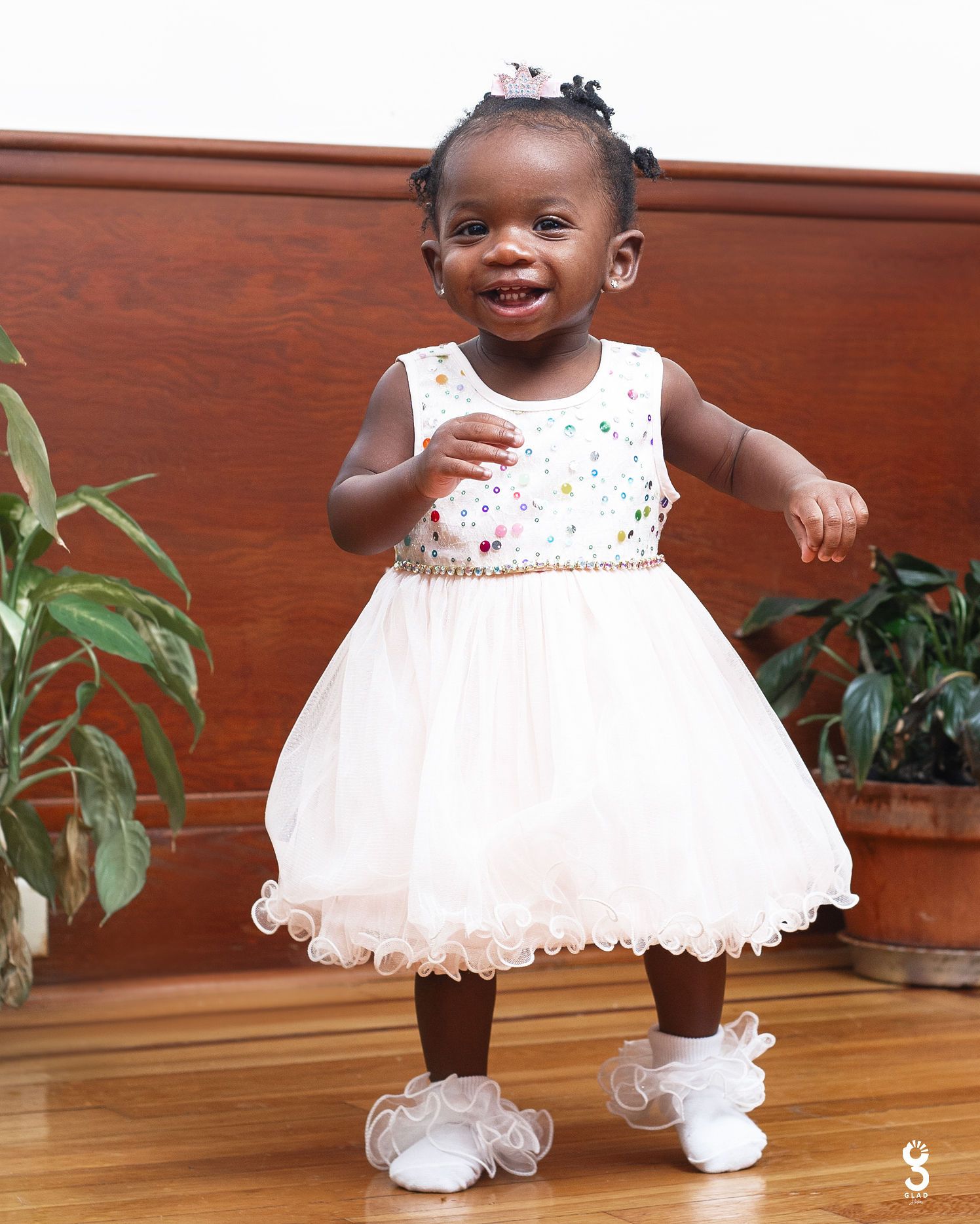 A toddler in a white party dress smiles and dances on a wooden floor.