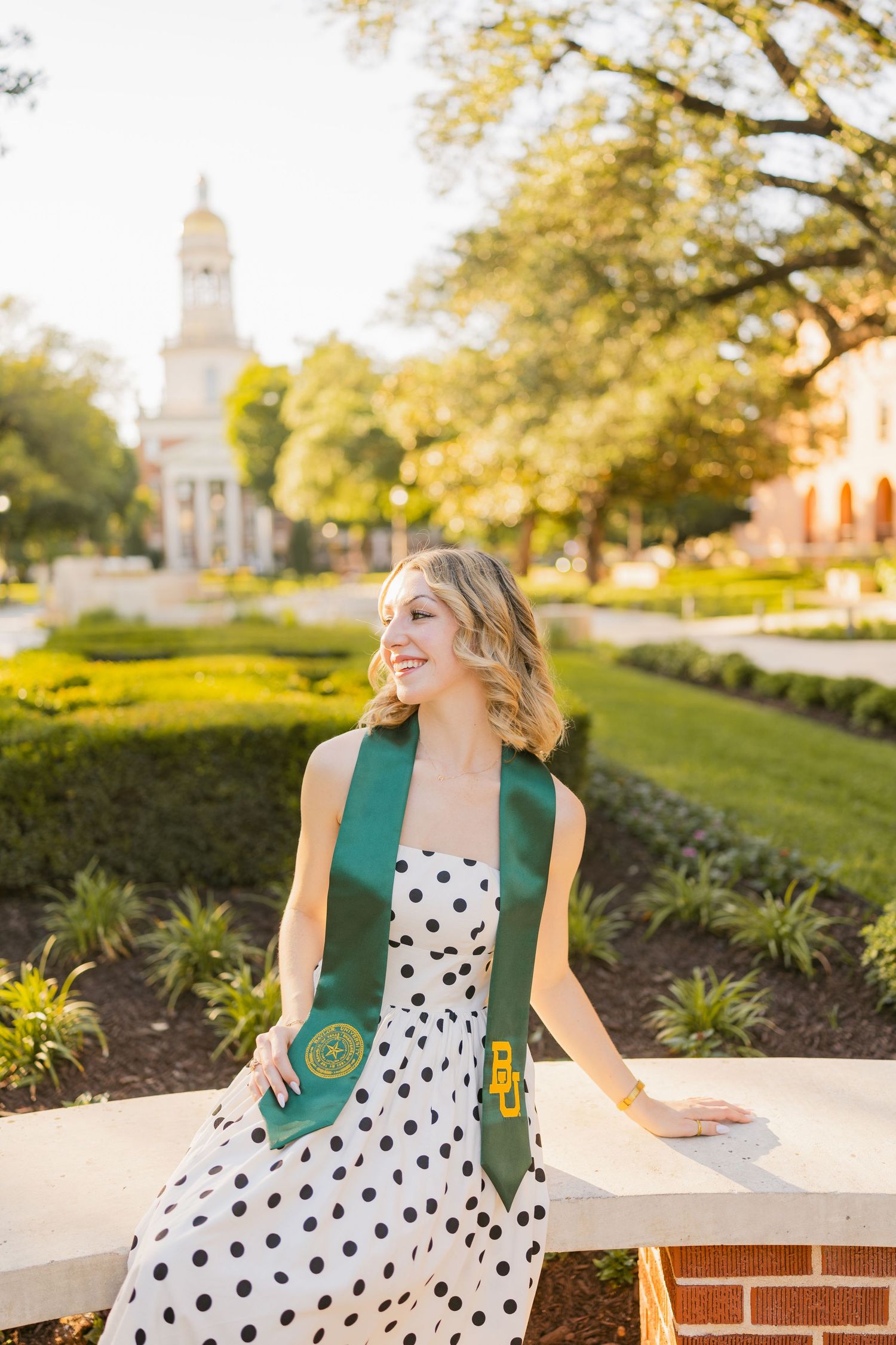 Blonde woman in polka dot dress with Baylor graduation stole seated on a brick wall outdoors.