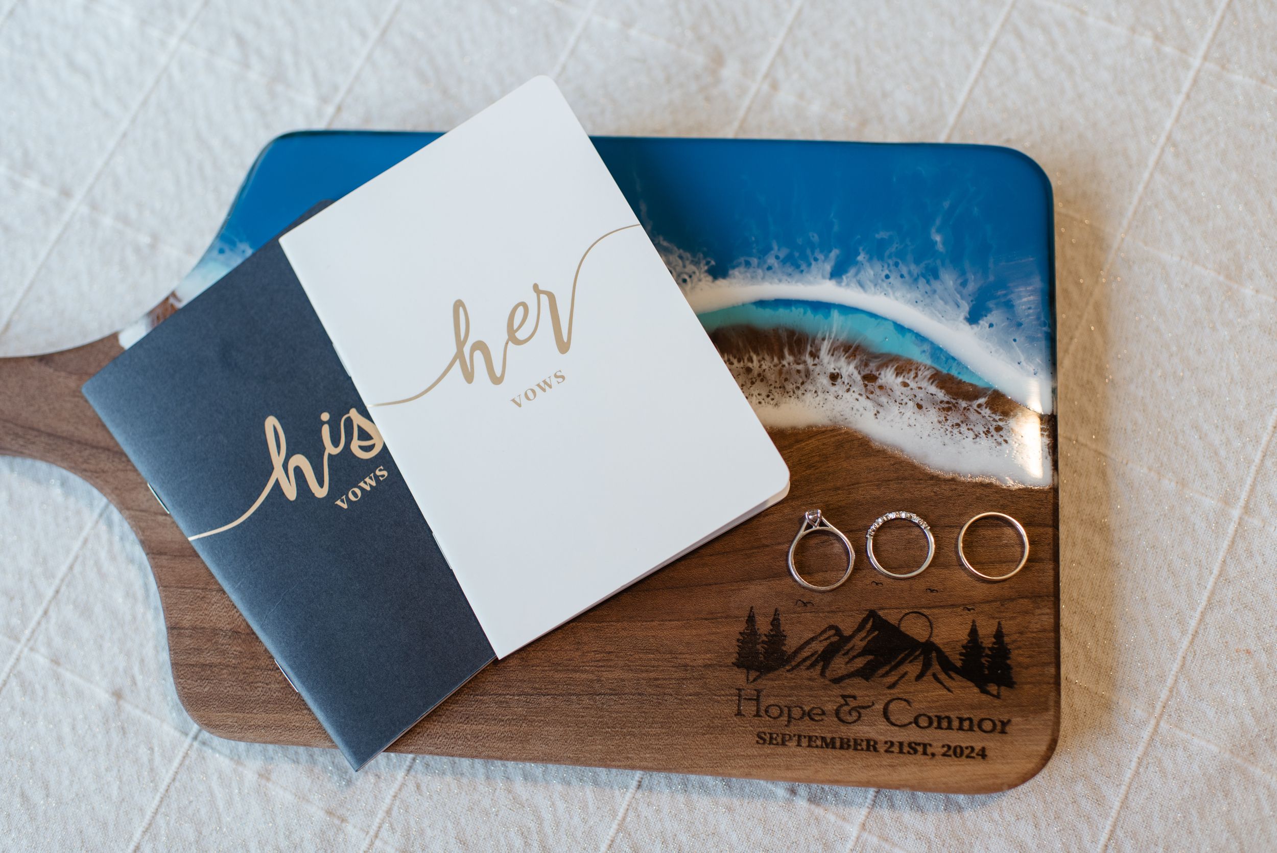 Two 'hello' wedding vow books and rings displayed on a wooden board with mountain landscape artwork decoration.