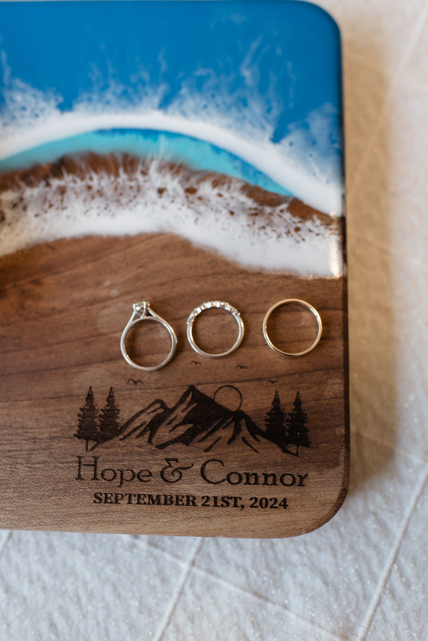 Three wedding rings arranged on a custom wooden board with mountain design and blue epoxy resin accent.