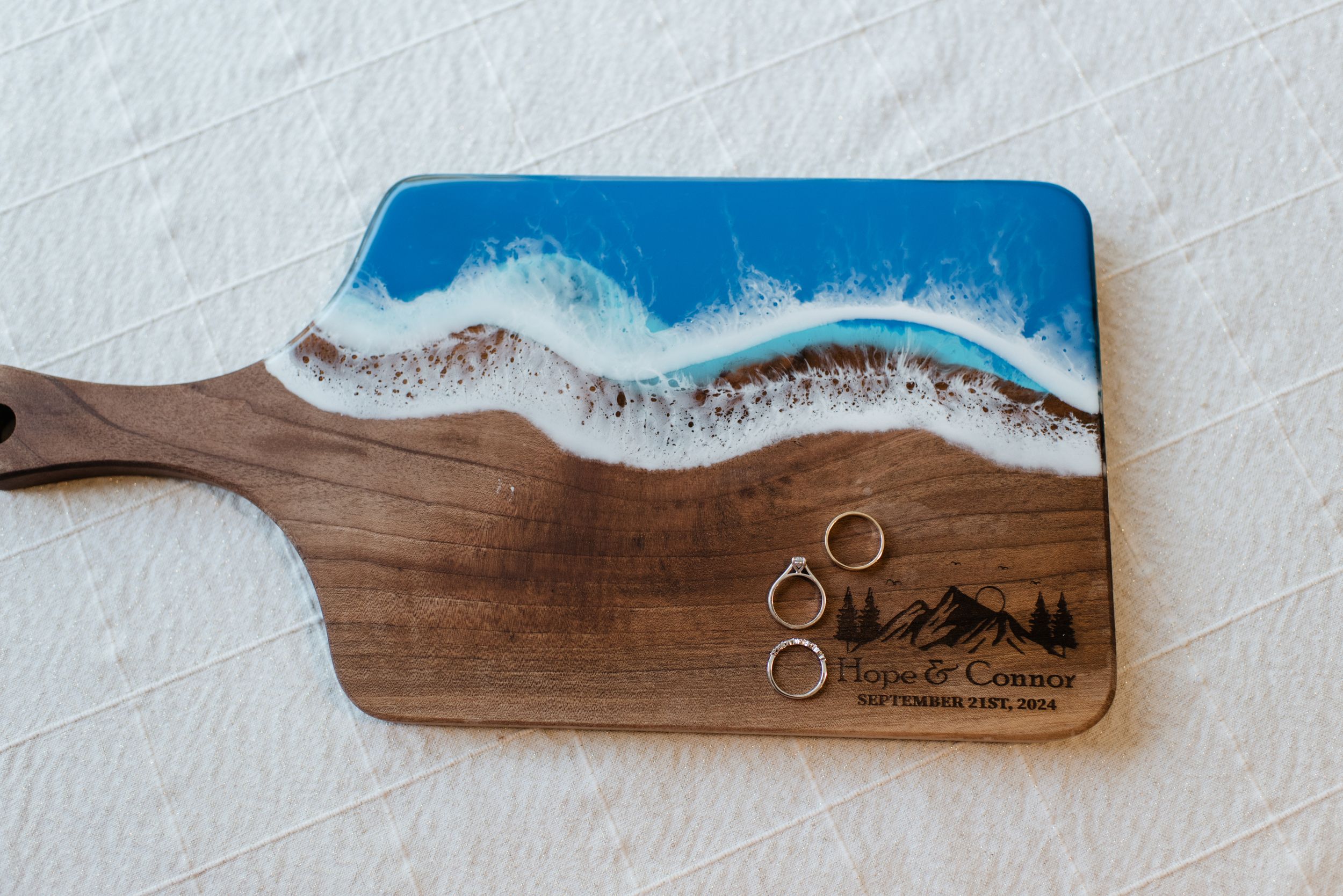 Wooden serving board decorated with blue and white resin ocean wave design and gold wedding rings on its surface.