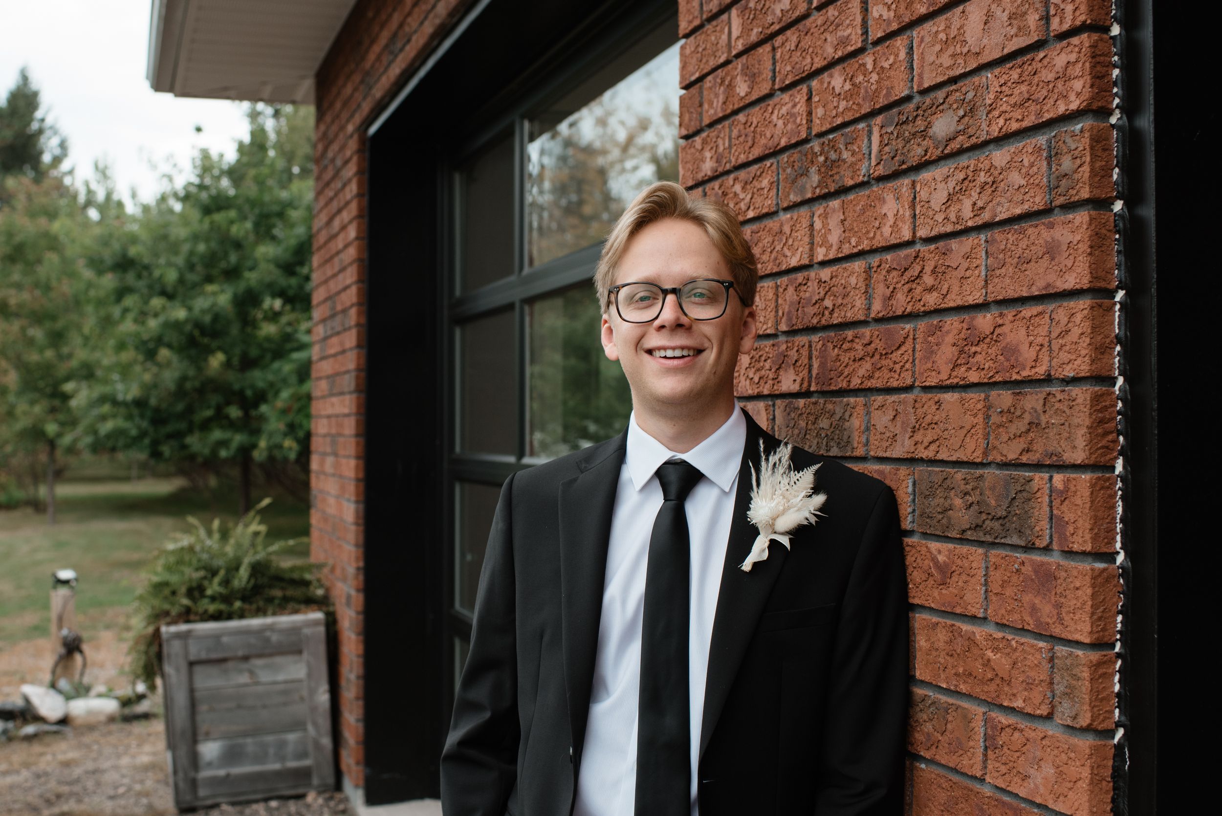A joyful person in a black suit and tie with a boutonniere smiles while standing against a brick wall.