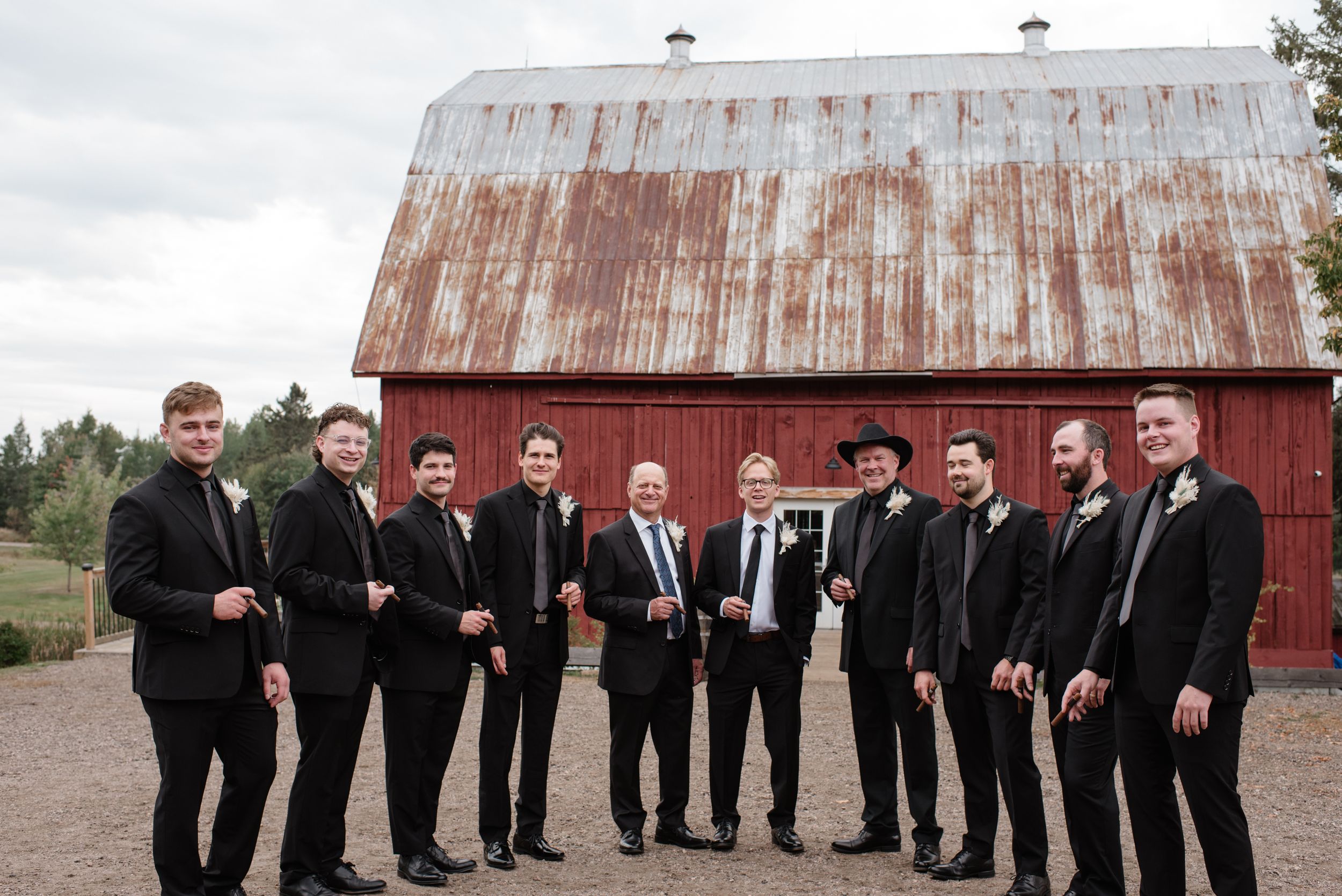 A group in formal black attire poses together in front of a rustic red barn with a weathered metal roof.