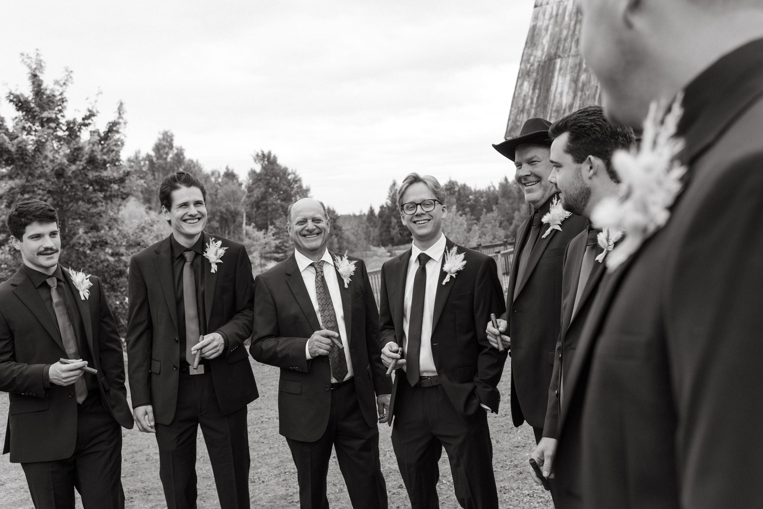 A cheerful group of groomsmen in black suits sharing a candid moment outdoors in black and white photography.