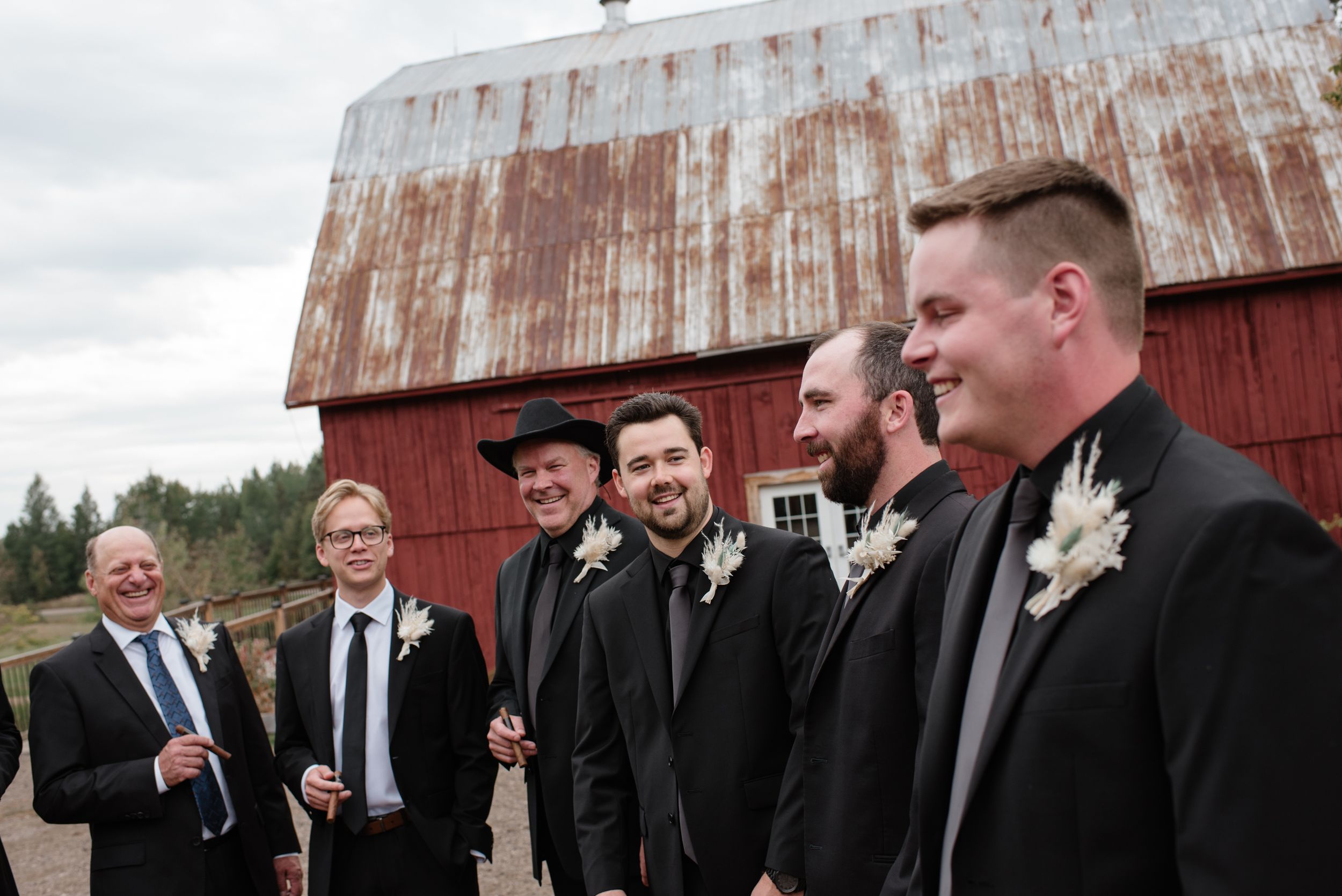 A group of men in formal black suits stand together near an old red barn on an overcast day.