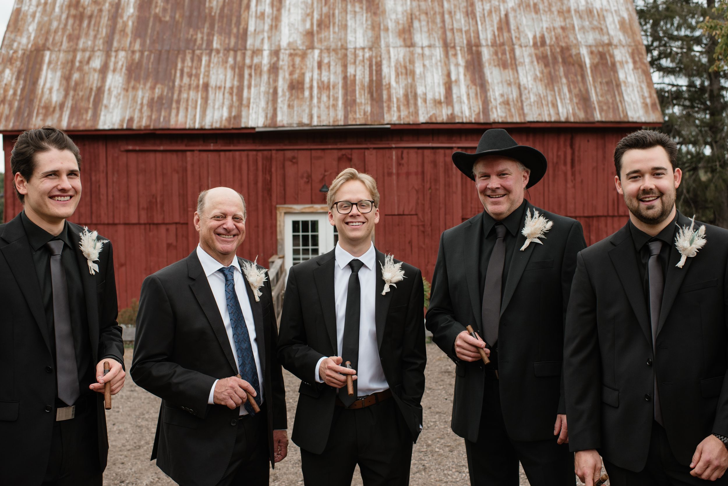 Group of groomsmen in black suits laughing and smiling together in front of a rustic red barn.