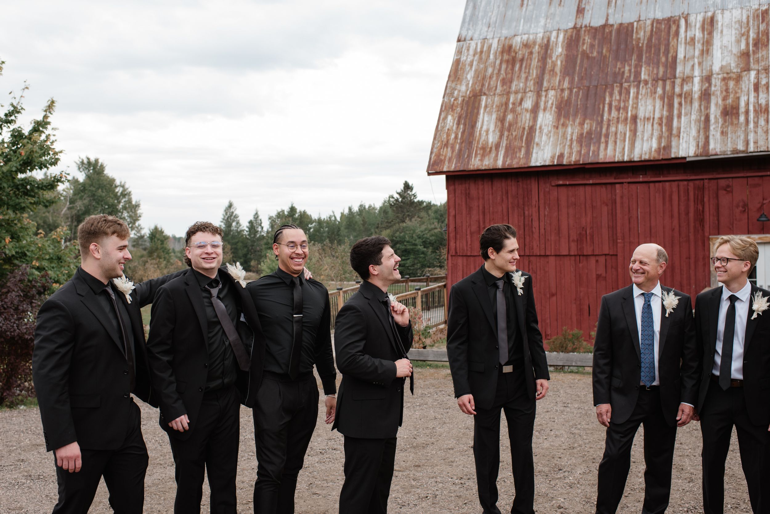 A group of well-dressed men in dark suits laughing together while standing outside a rustic red barn in the countryside.