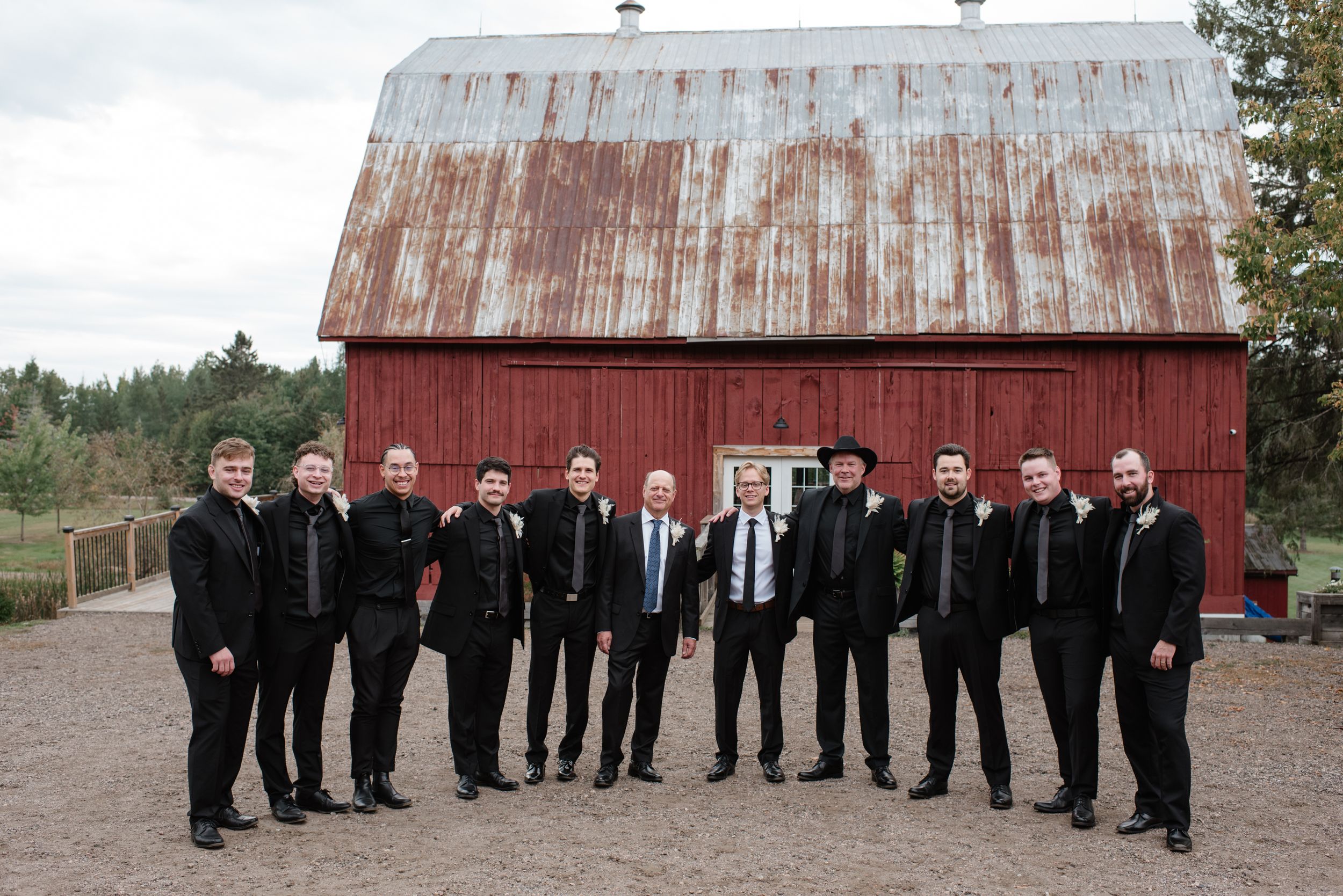A group of formally dressed people in black suits stand together for a photo in front of a rustic red barn.