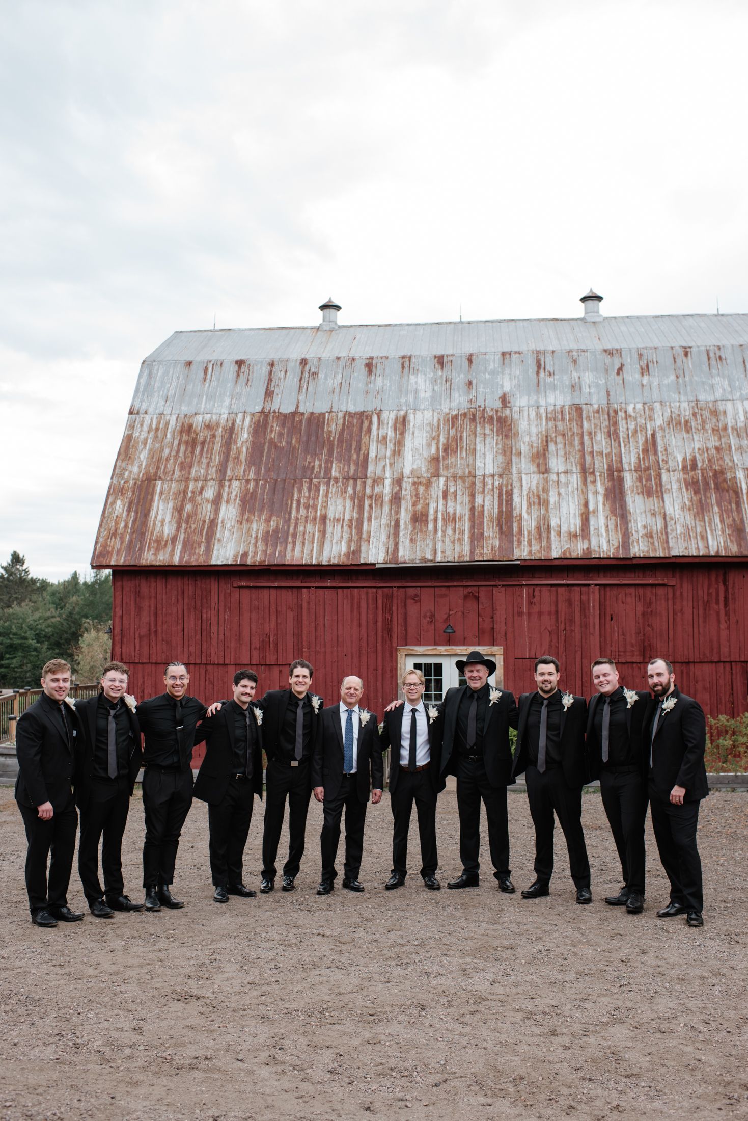 Groomsmen lined up in black suits standing in front of a rustic red barn with weathered metal roof.