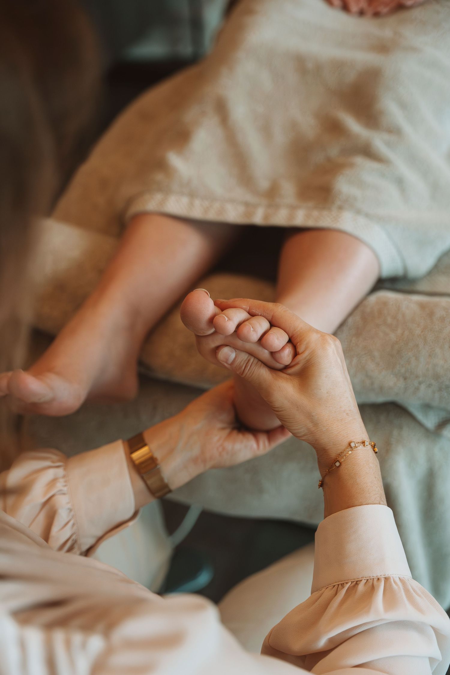 A gentle foot massage being given on a couch with soft lighting and delicate gold bracelets visible on the masseur's wrists.
