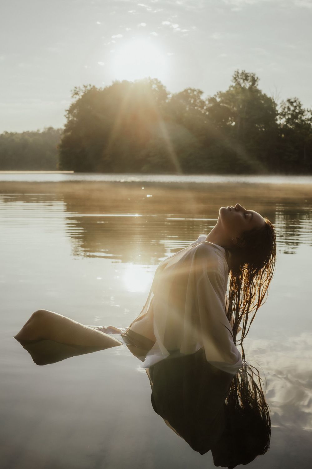 Silhouette of a person floating in tranquil lake water at sunset with sunbeams streaming through trees.