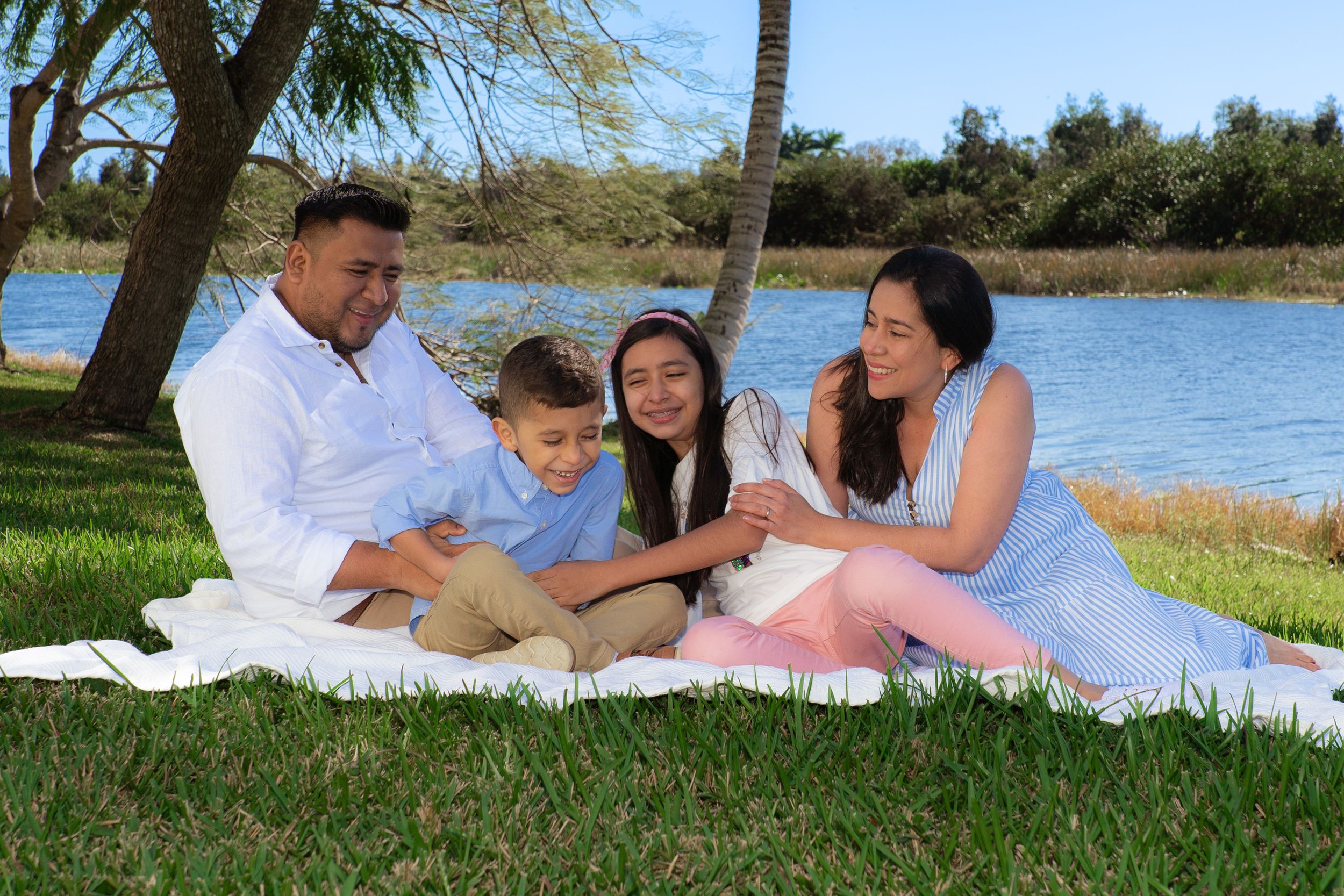 Family of four sits on a blanket by a lake, sharing a warm moment together.