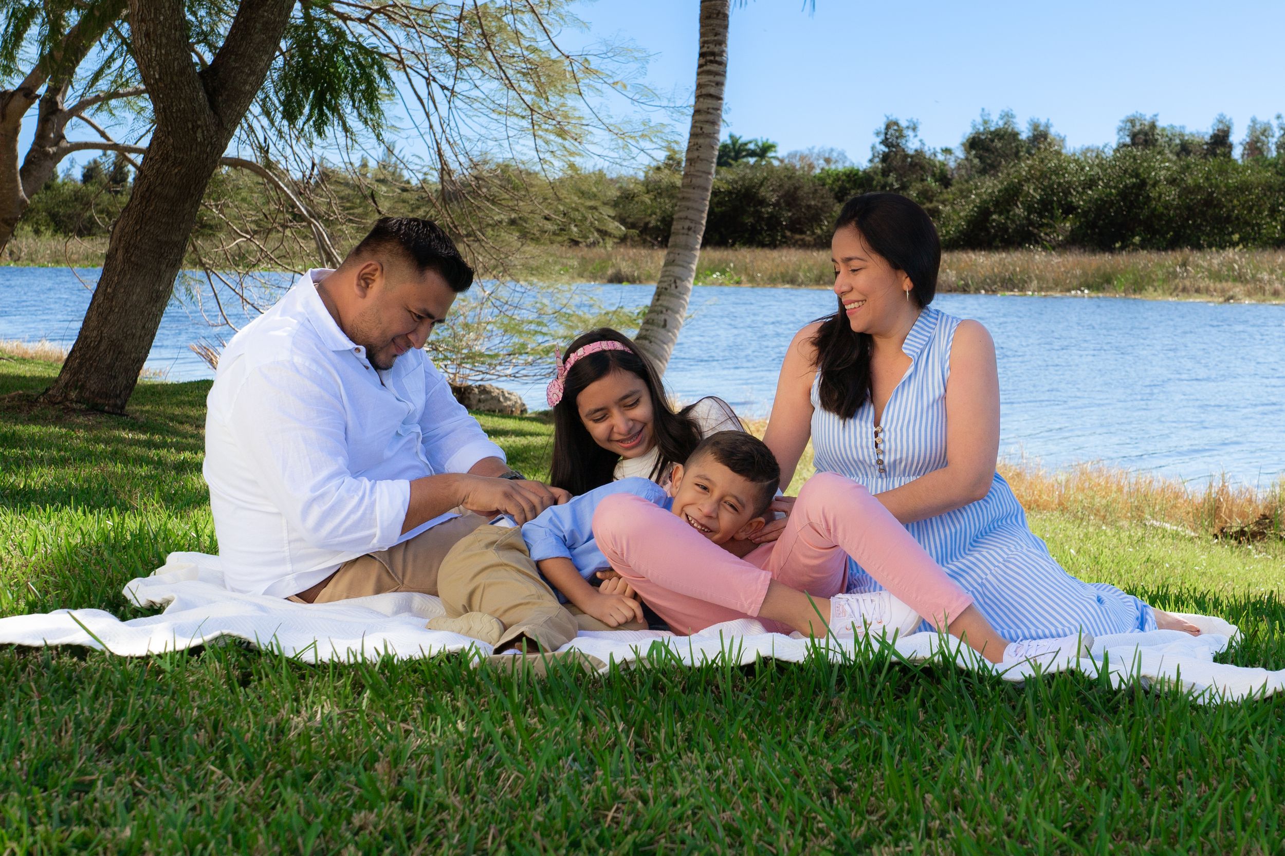 Family relaxes on a white blanket by a serene lake, children leaning on parents.