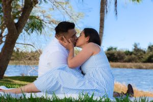 Couple sharing a romantic kiss while sitting on a blanket by a serene lake surrounded by lush greenery.