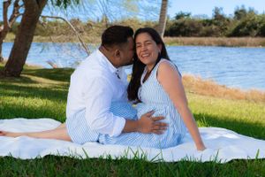 Happy pregnant couple sitting on a white blanket by a lake, sharing a joyful embrace outdoors.