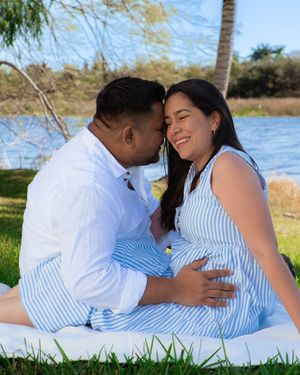 Expecting couple sitting on a blanket by a lake, man kissing woman's cheek while cradling her baby bump.