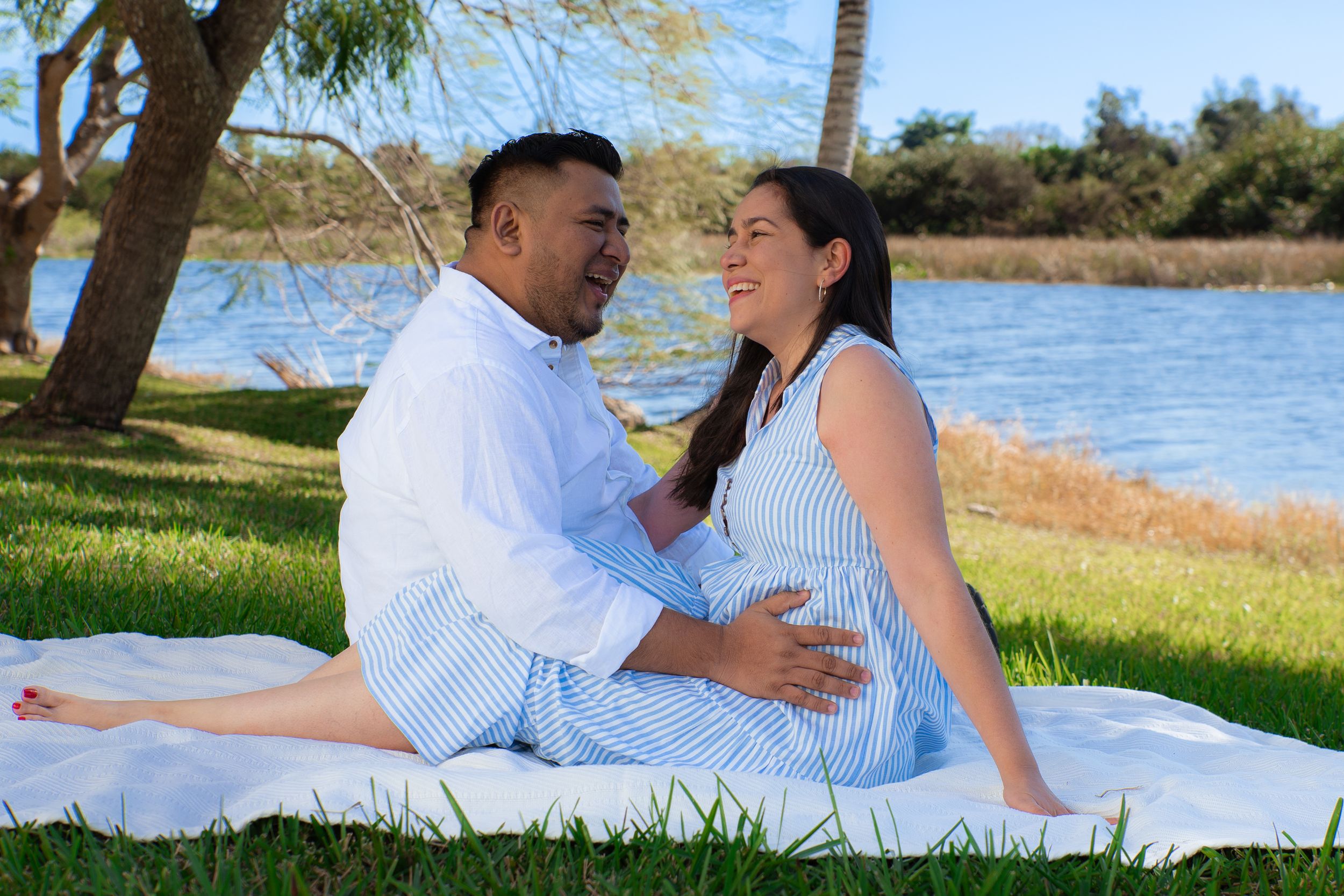 Smiling couple sitting on a blanket by a lake, tenderly holding the woman's pregnant belly together.