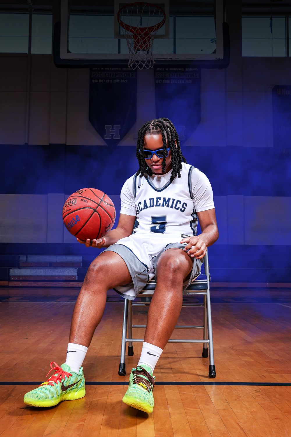 Basketball player wearing white Academics jersey sits on chair with neon yellow sneakers on wooden court.