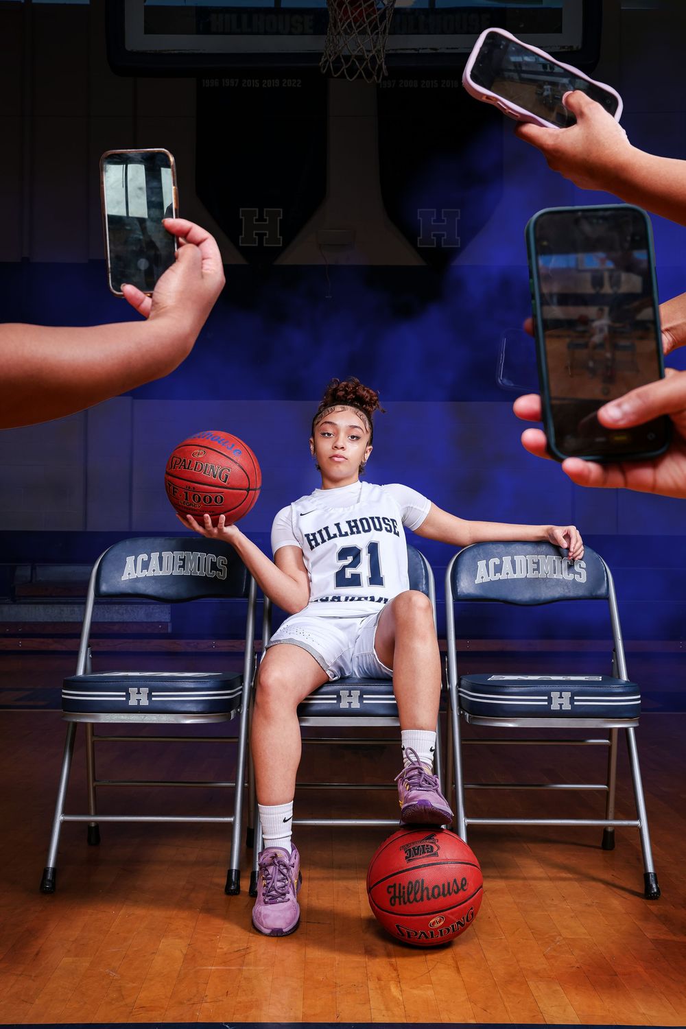 Basketball player sits between folding chairs on court while phones capture photos in dramatic lighting setup.