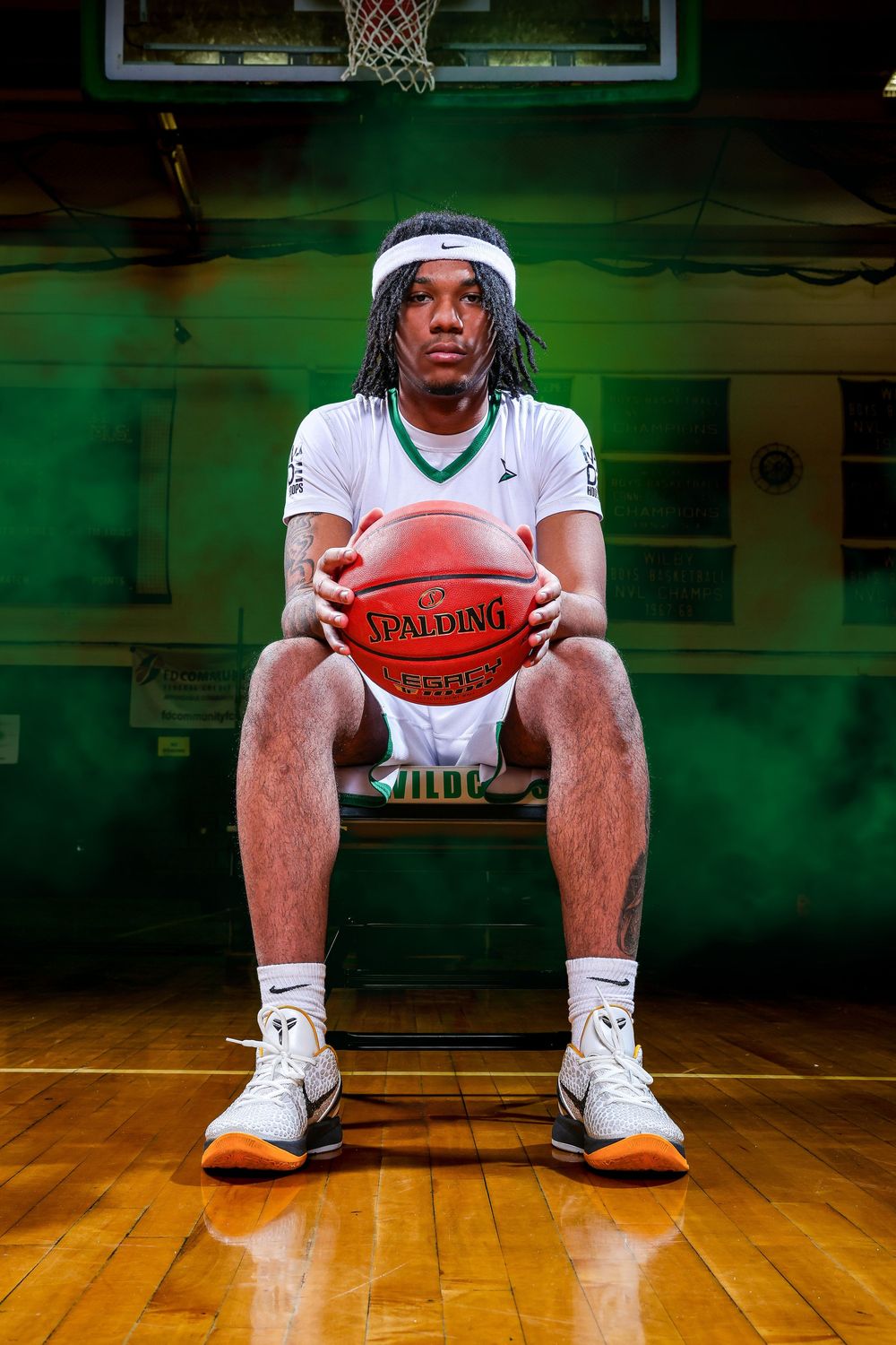 Basketball player sits on wooden court floor holding ball with dramatic green lighting and basketball hoop in background.