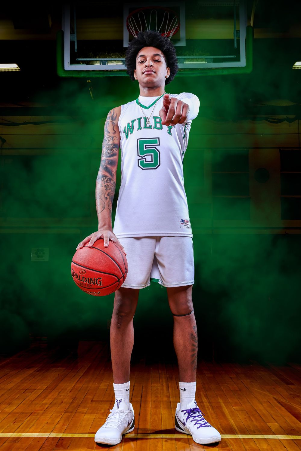 Basketball player in white jersey with number 5 poses on wooden court with green backdrop.
