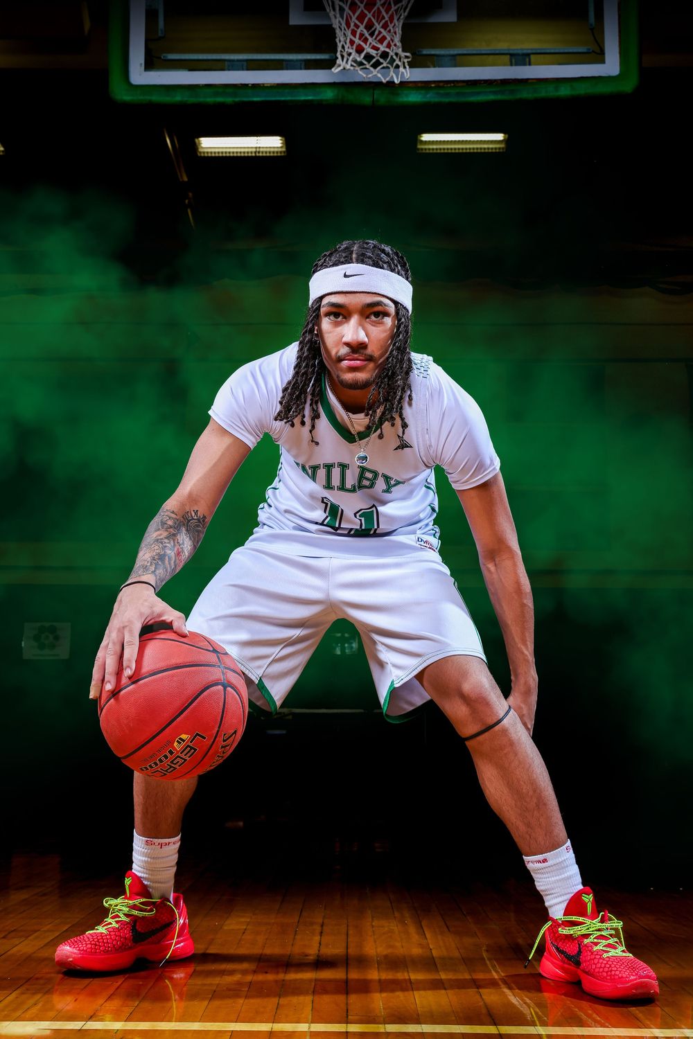 Basketball player in white Wiley uniform dribbles in athletic stance on court with green backdrop and hoop visible.
