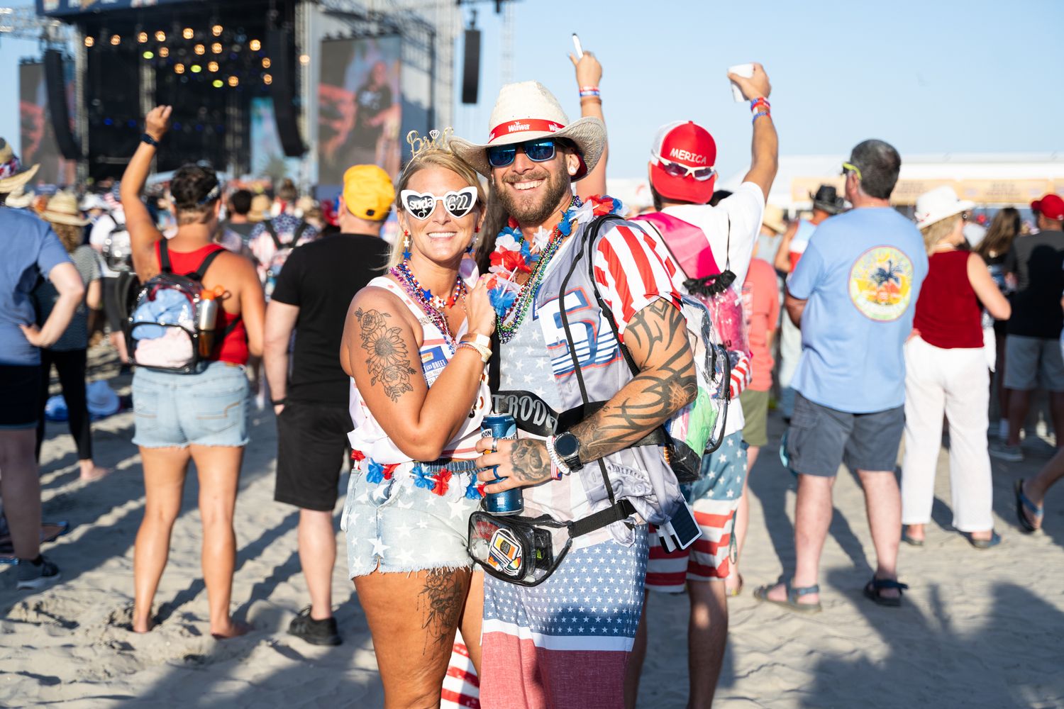 A couple enjoying a music festival, smiling and dancing together on the sandy ground under a bright blue sky.