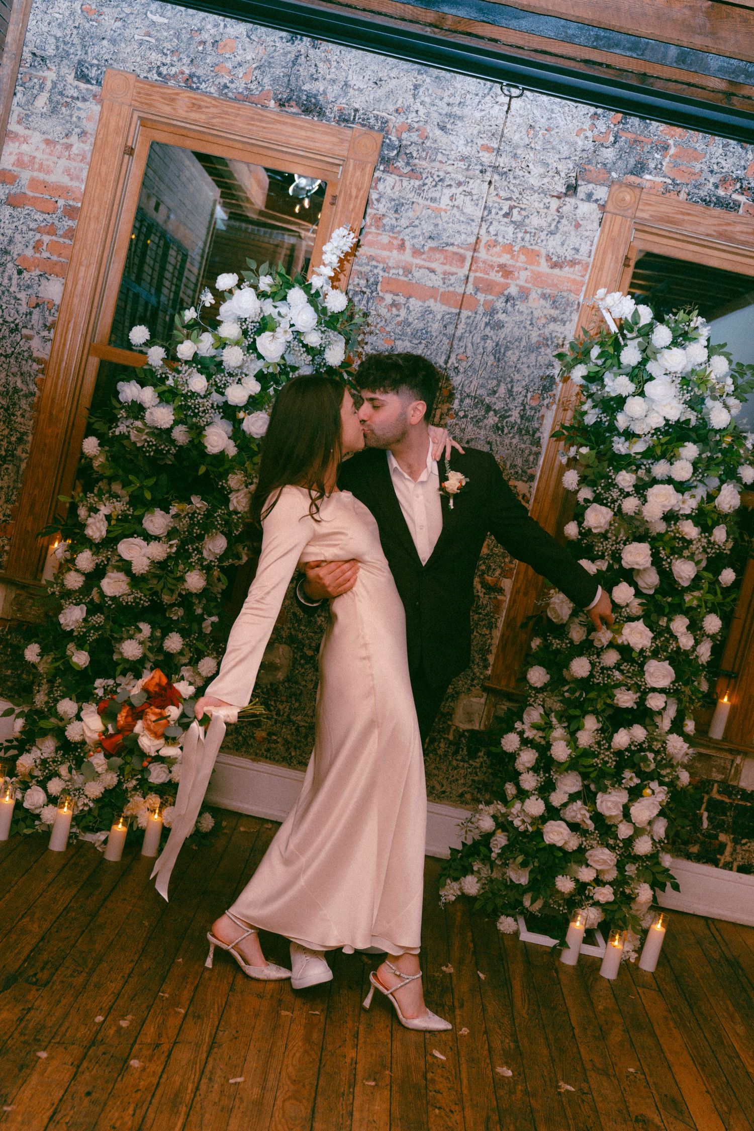Couple sharing a romantic kiss at wedding altar adorned with white floral arrangements and candles in rustic brick venue.