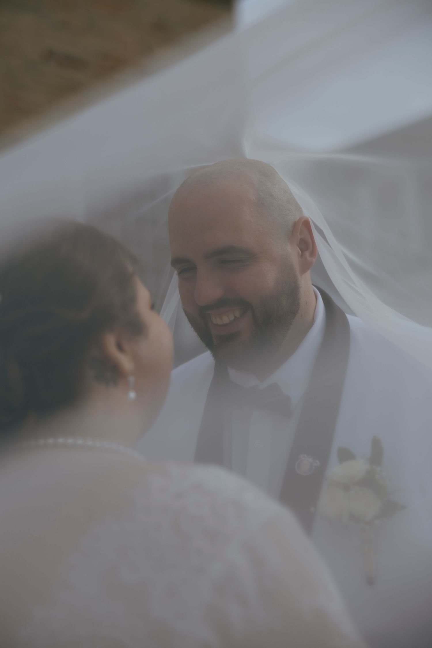 Bride and groom smiling under wedding veil, groom in white suit with boutonniere, romantic outdoor ceremony moment.