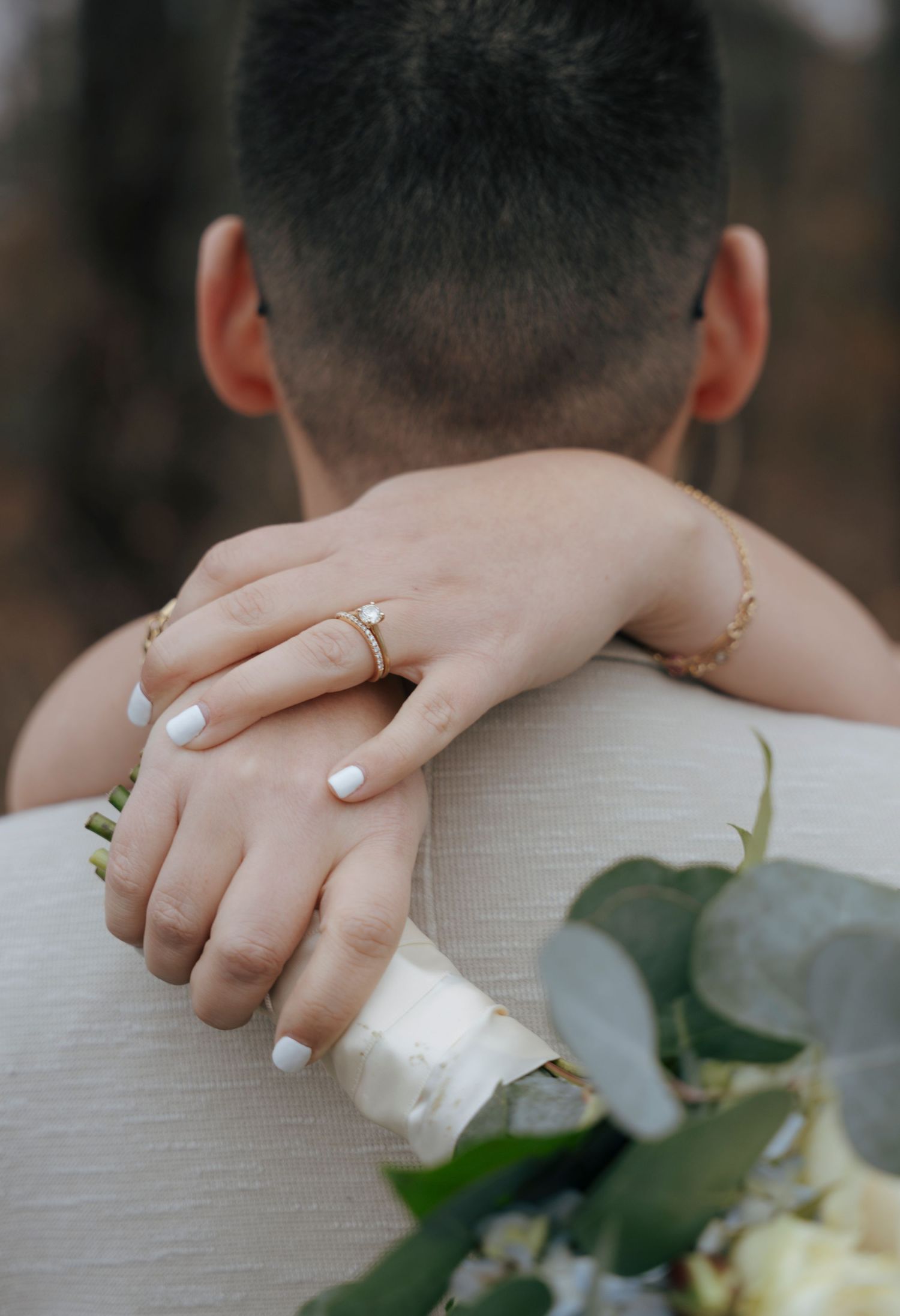 Bride resting hands on groom's shoulder, displaying diamond engagement ring and white manicure while holding a floral bouquet.
