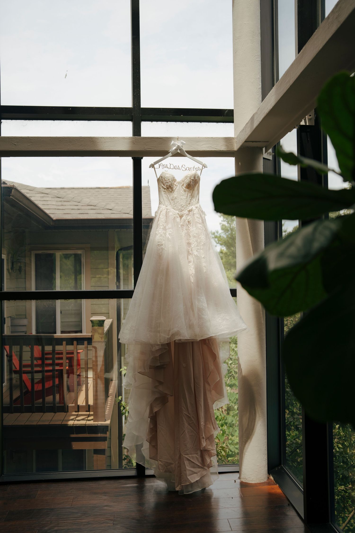 Elegant strapless wedding dress with lace bodice and tulle skirt hanging on a decorative hanger in a sunlit window.