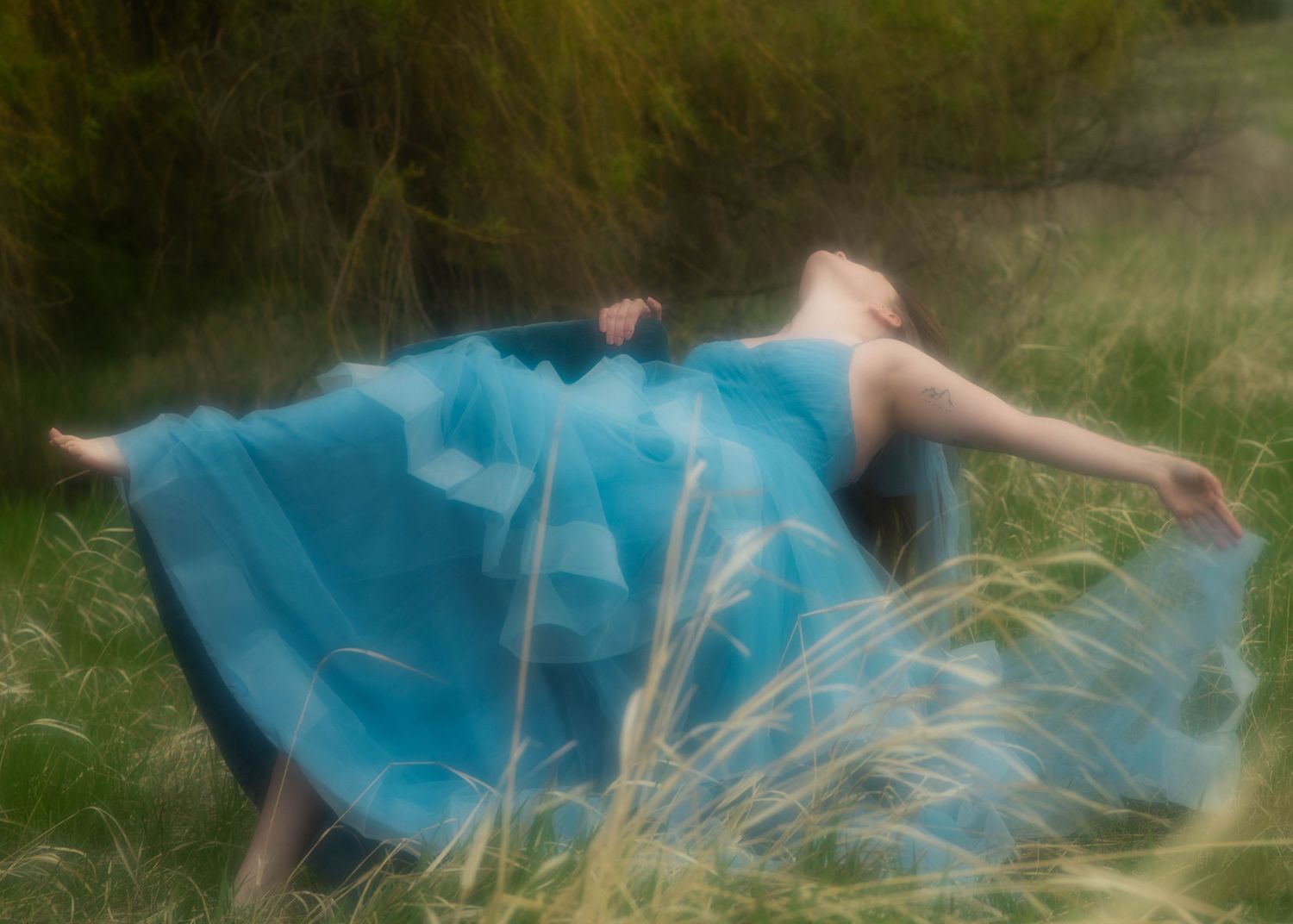 Woman in flowing turquoise gown lying in tall green grass with arms outstretched in dreamy outdoor setting.