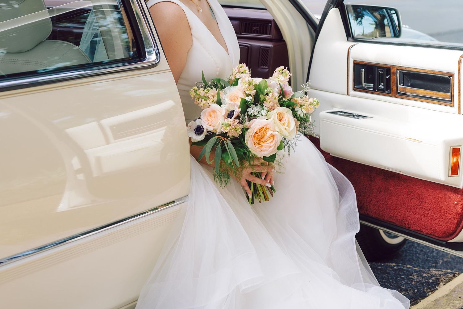 Beautiful peach and cream bridal bouquet rests on a white tulle gown inside a classic car with beige and red interior.