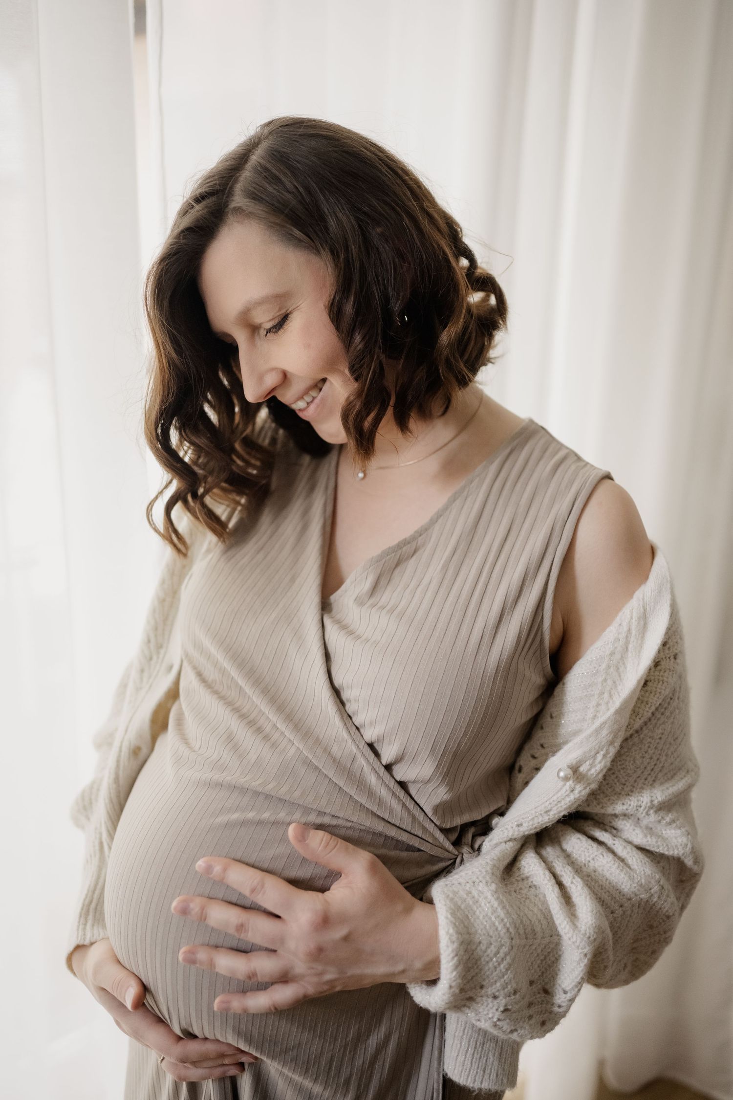 Expectant mother in soft beige dress and cardigan gently cradles her baby bump by window in natural light.