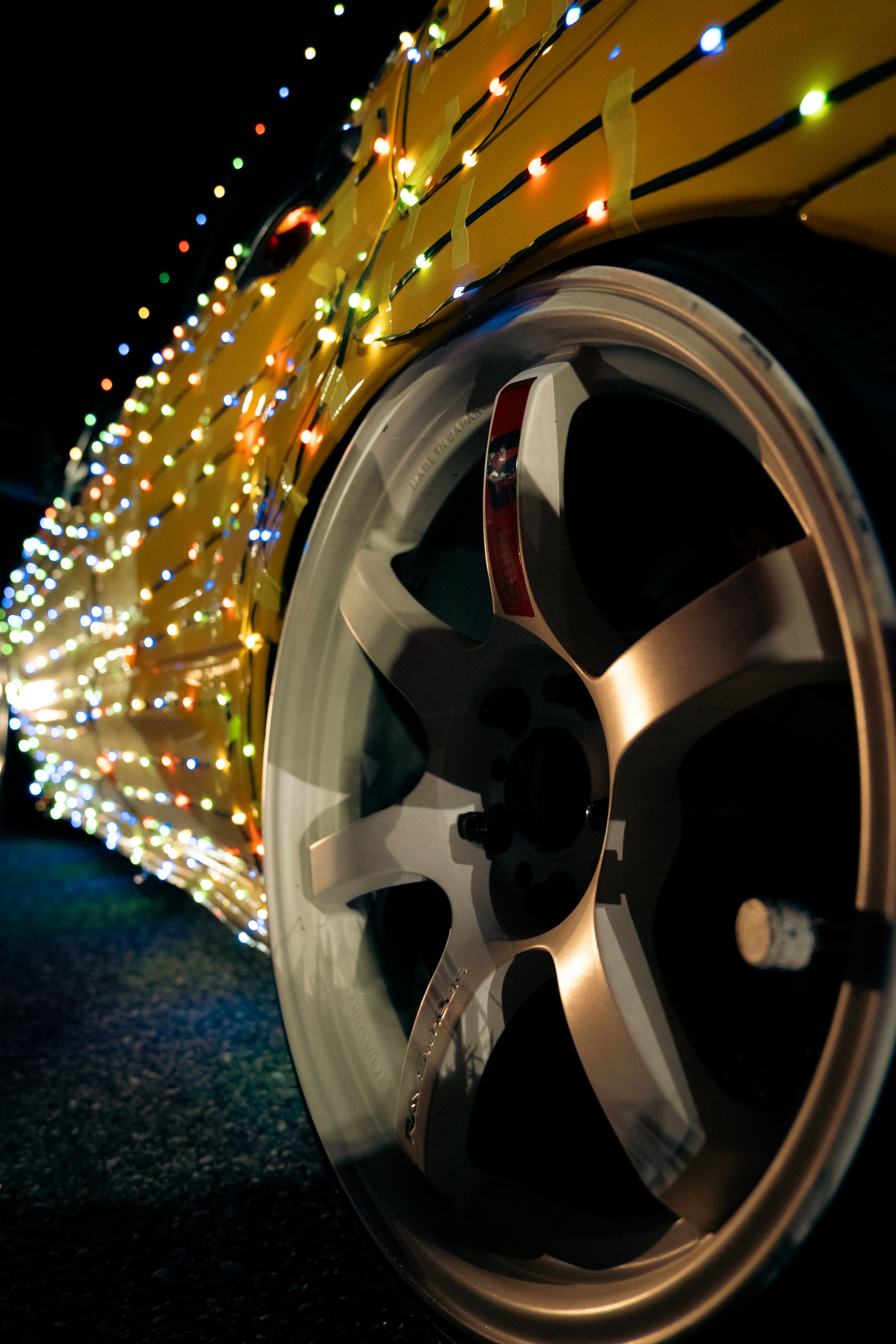 Close-up of a chrome alloy wheel on a yellow car decorated with multicolored Christmas lights.