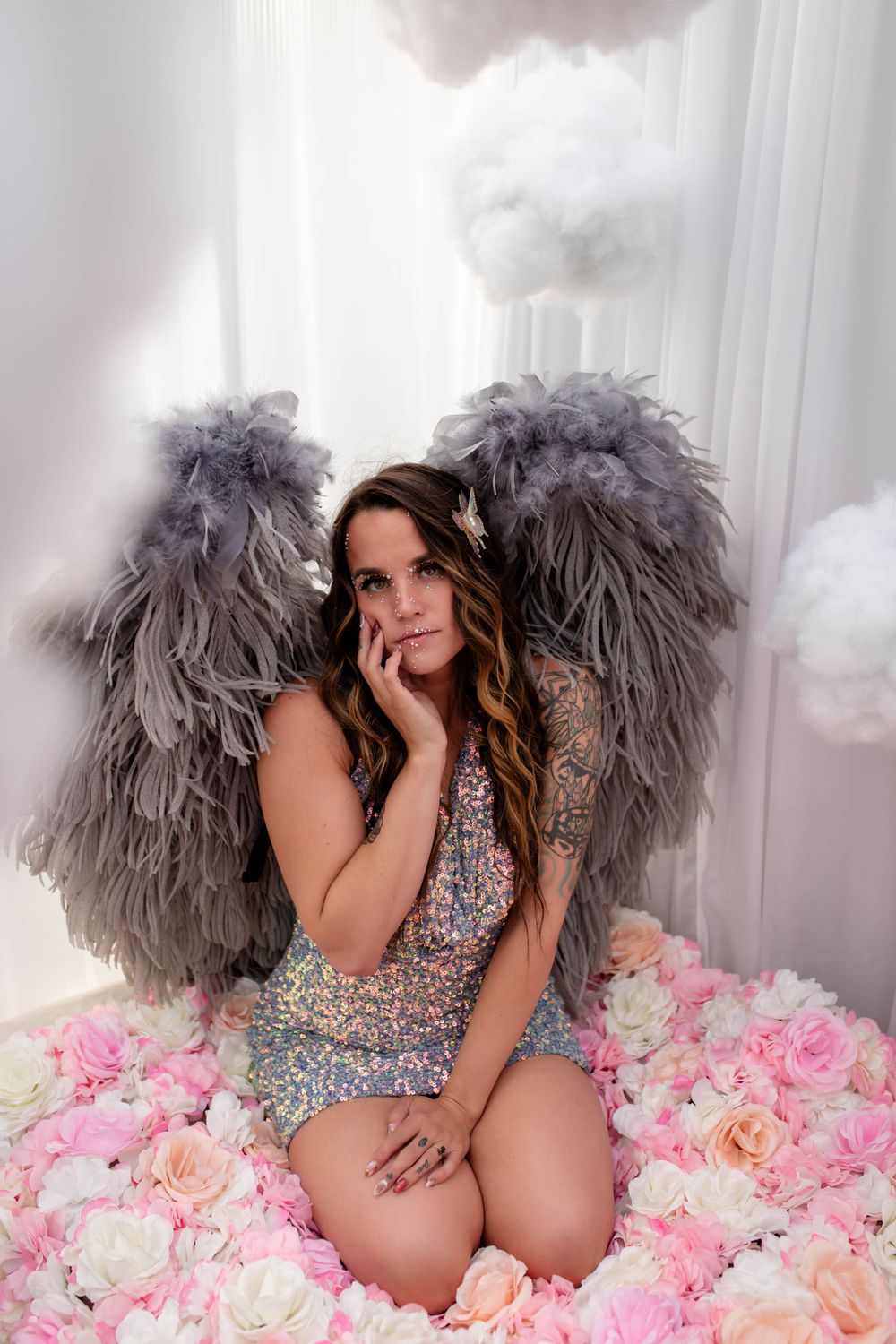 Fantasy photoshoot with gray feathered wings and sparkly dress amid pink and white roses.